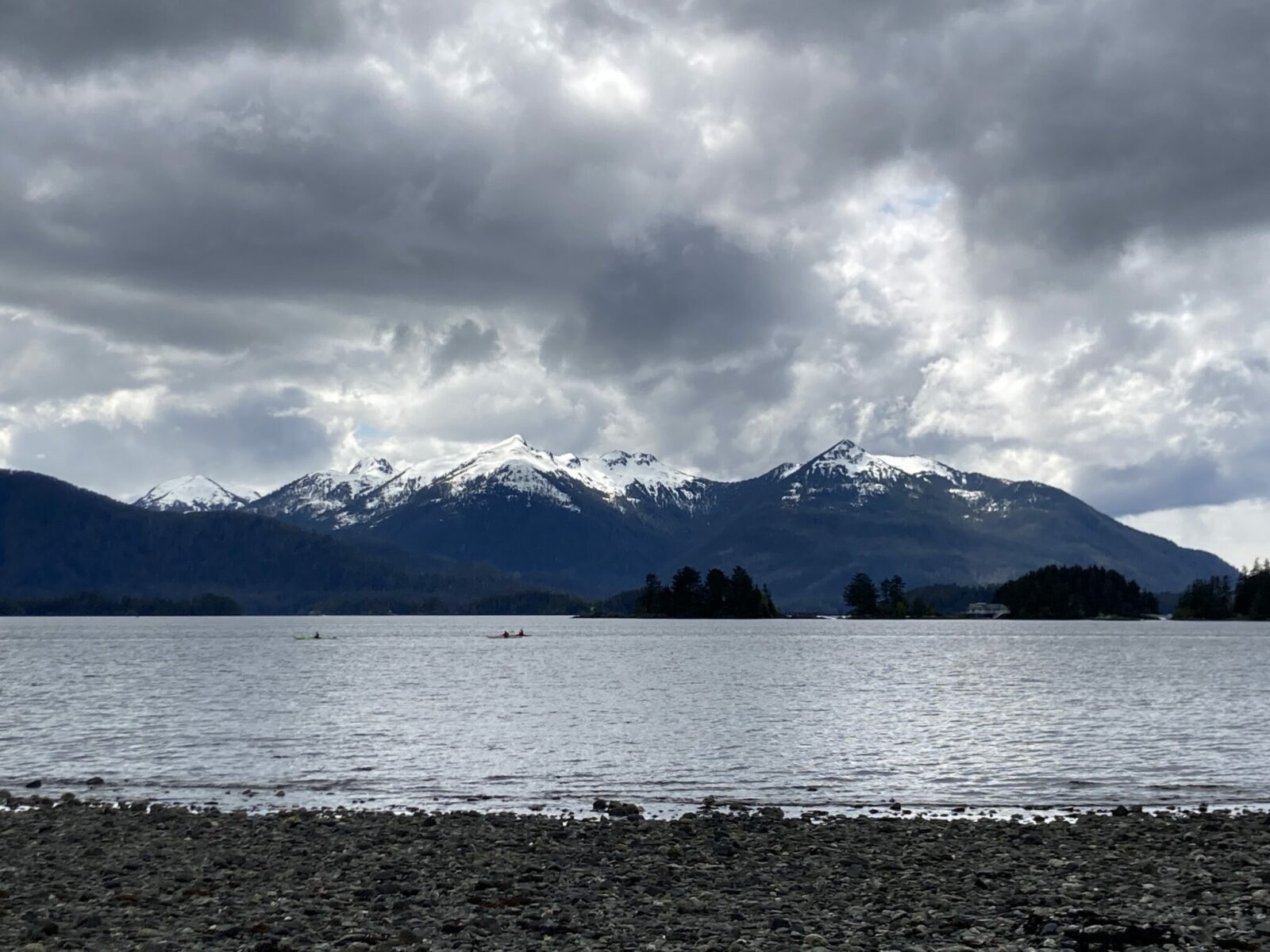 Three kayakers paddling off the coast of Sitka, Alaska. There is a rocky beach in the foreground and snow capped mountains and forested islands in the distance. The sky is overcast