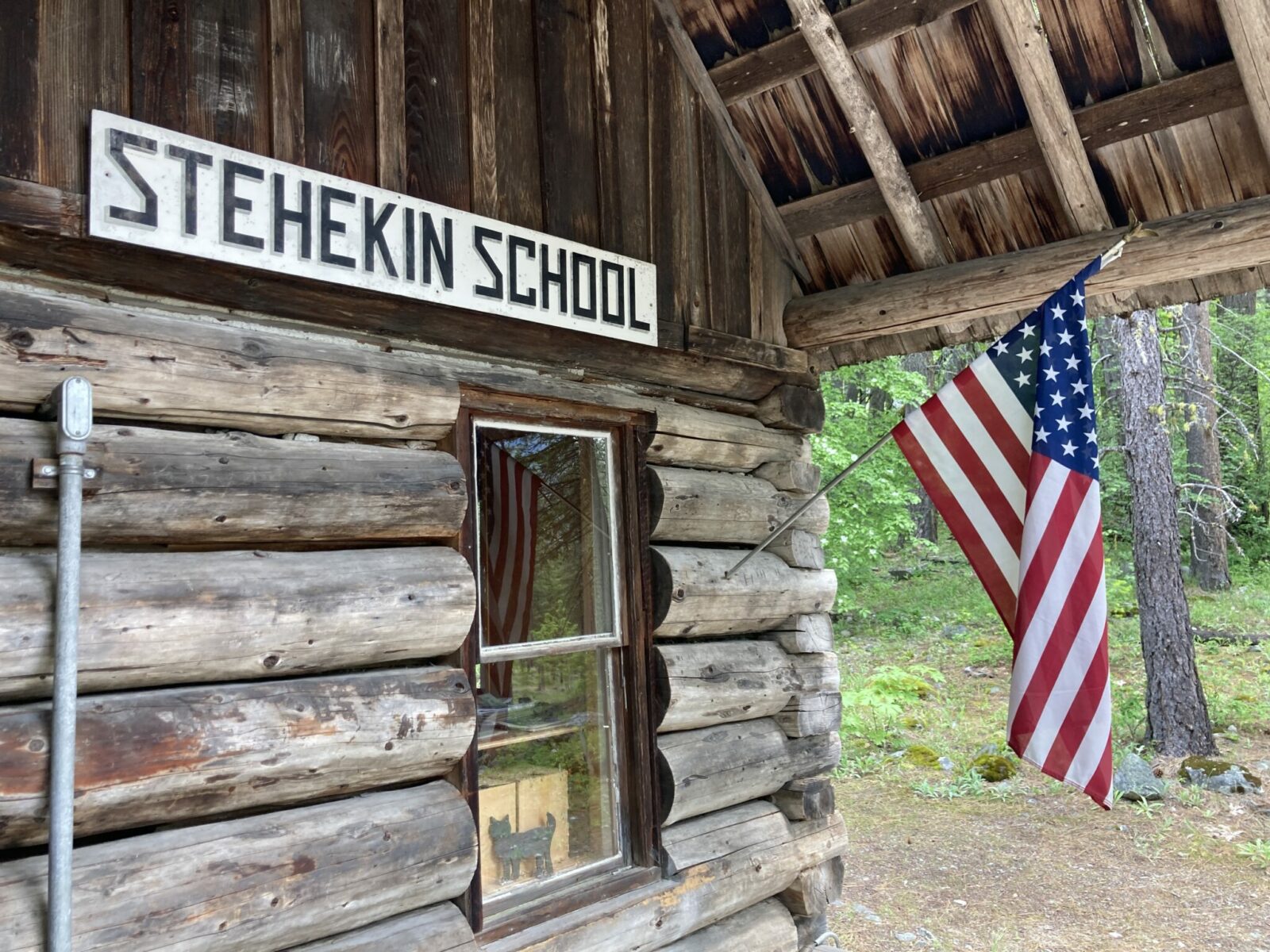 The side of a historic log building with an American flag. A sign over the window says Stehekin School