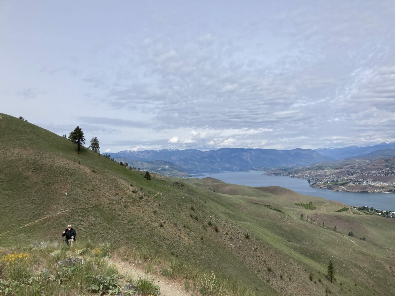 A hiker on a dirt trail on a hill above Lake Chelan. Below you can see the trail winding up the hill as well as the town, the lake and distant mountains