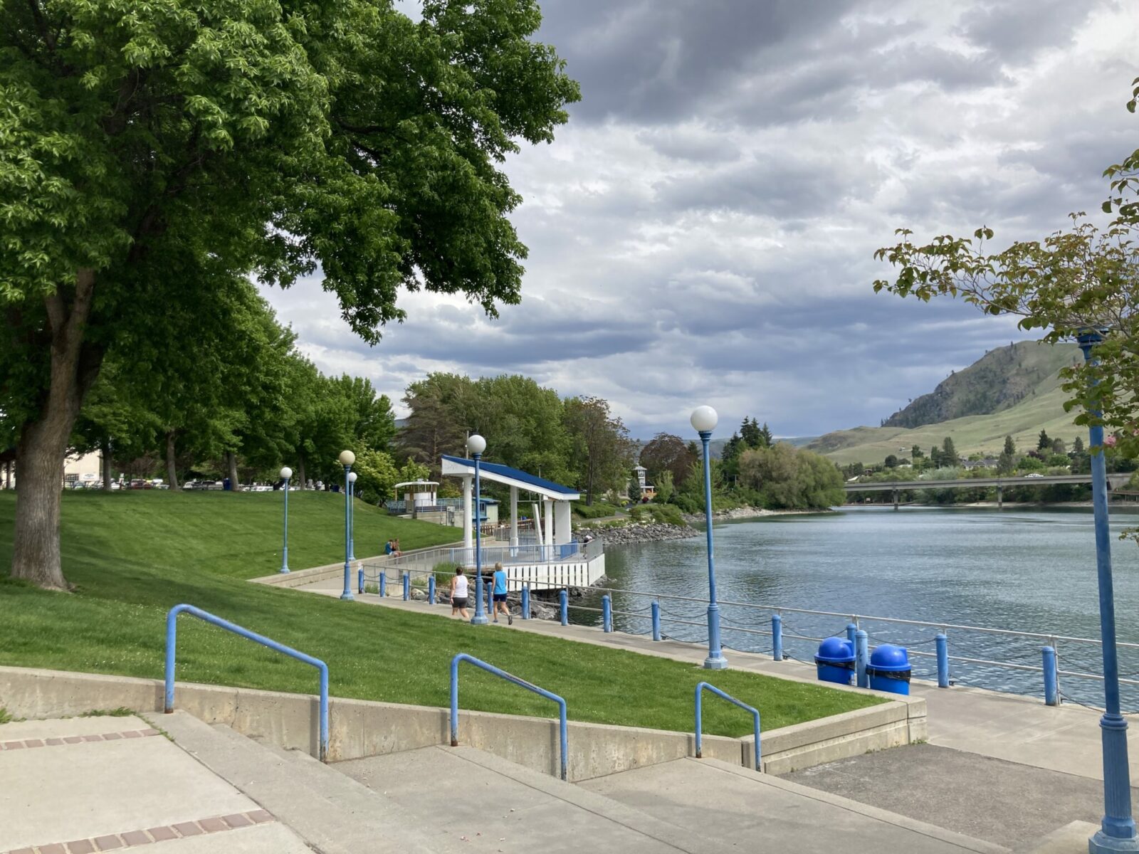 A city park with a paved trail and blue lightpoles and handrails. The park is lined with trees and next to a river in Lake Chelan