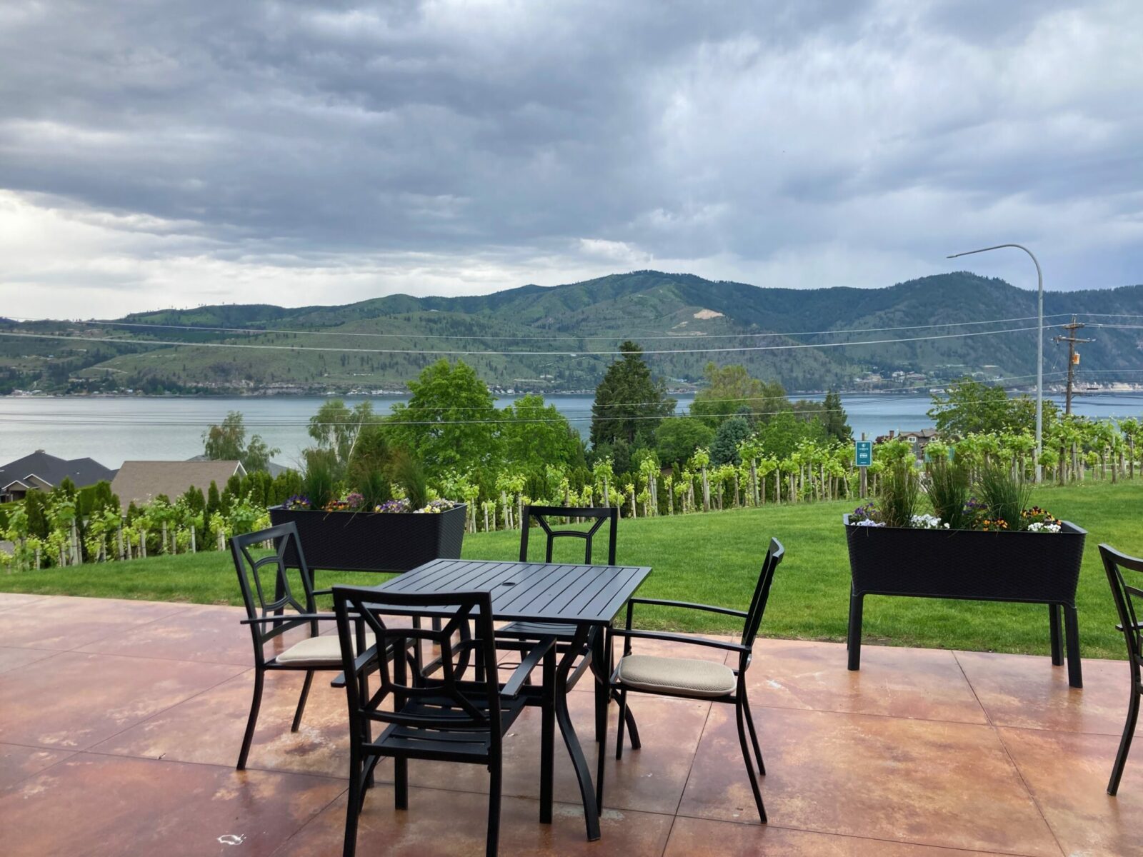 a patio with tables and chairs next to a lawn and a vineyard with a view down to trees, homes and Lake Chelan.