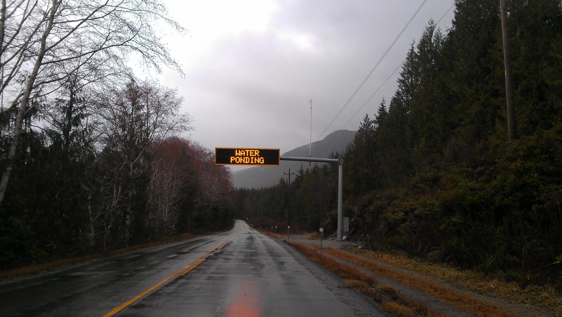 A two lane highway on an overcast and rainy winter day between Nanaimo and Tofino. A sign over the road says "Water ponding"