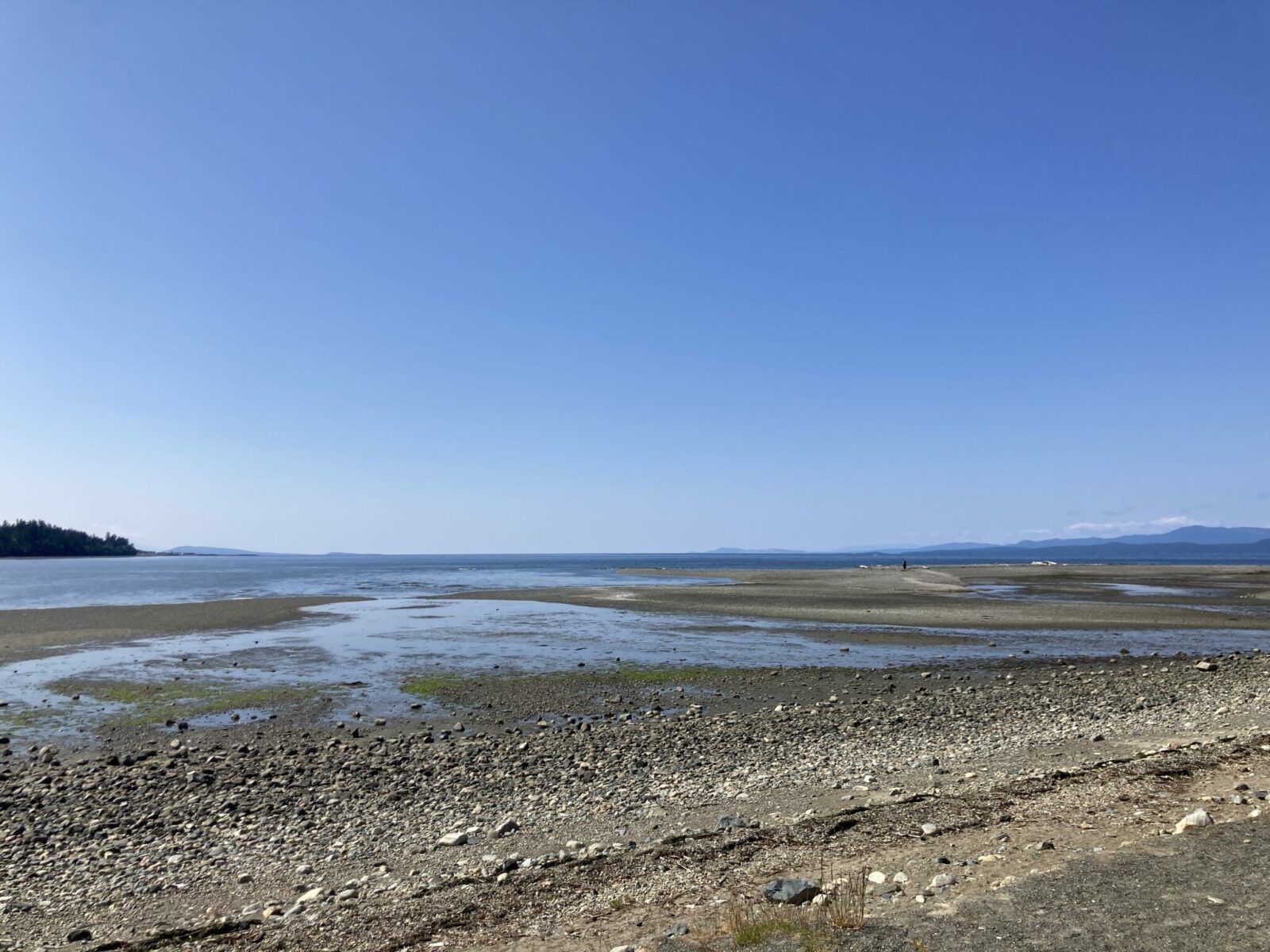 A gravel beach at low tide with distant forested mountains.