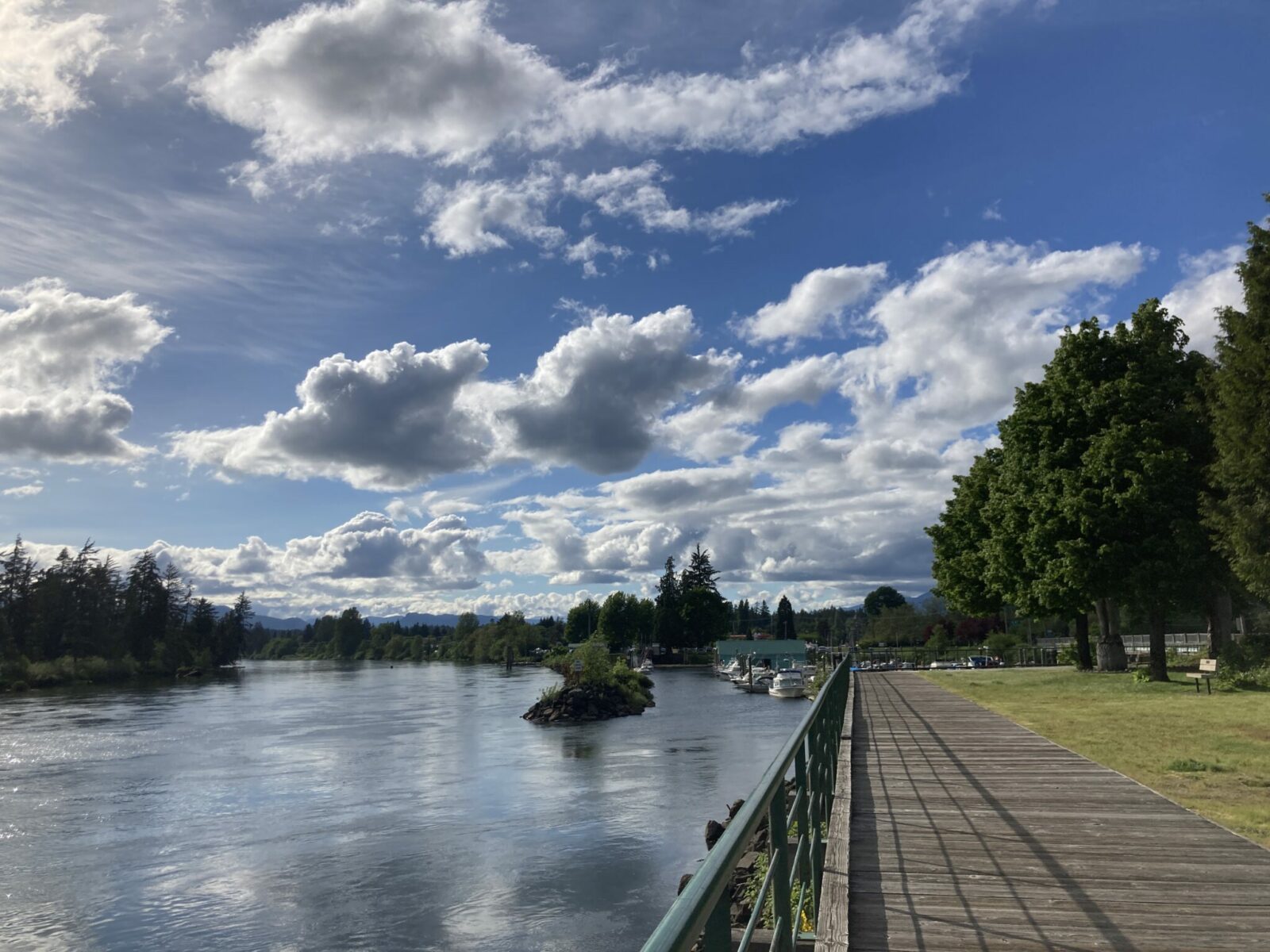 A wooden boardwalk along a river with grass and trees to the right and a marina directly ahead.