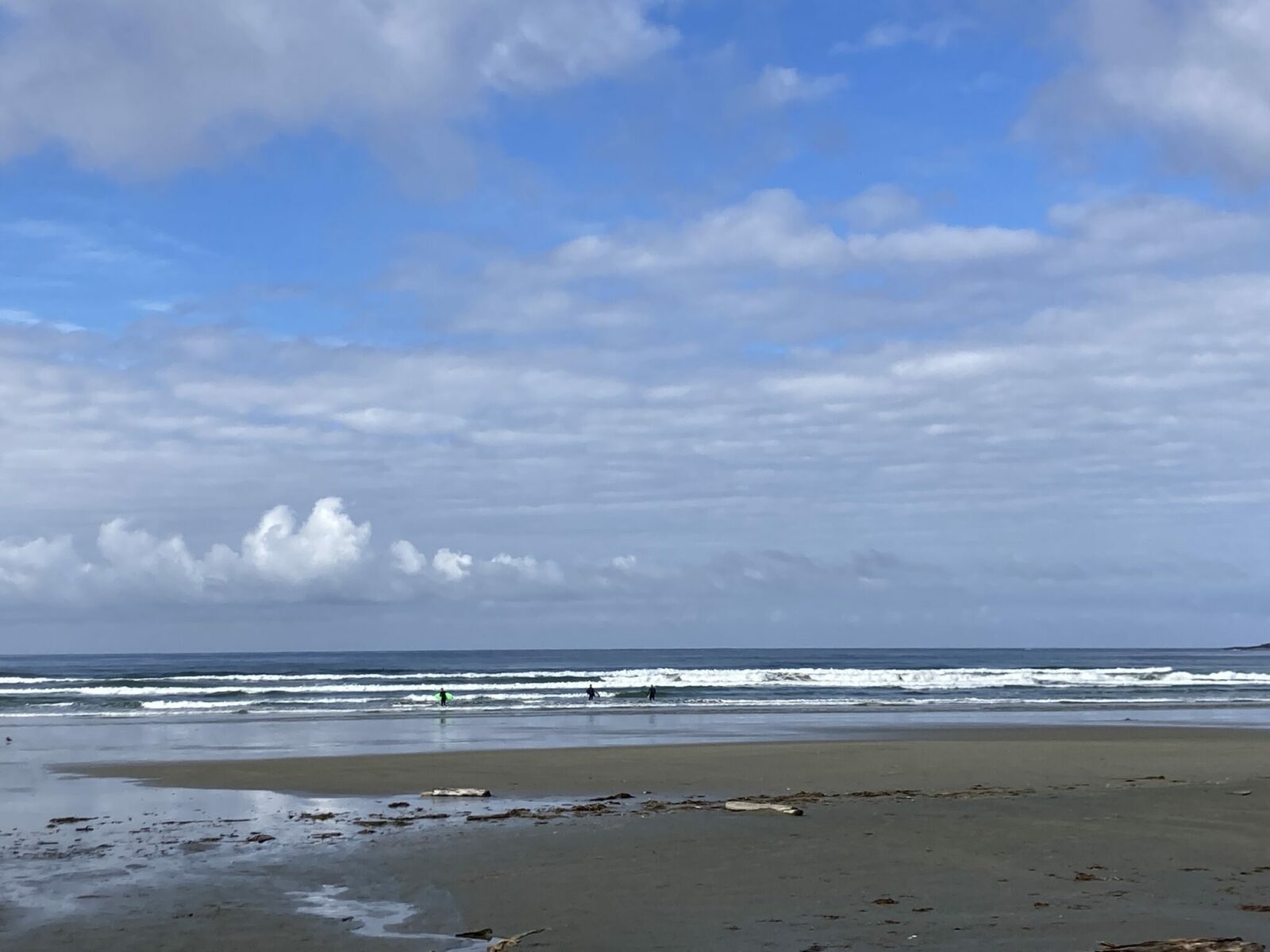 A sandy beach with ocean waves crashing on Long Beach in Pacific Rim National Park near Tofino. There are clouds offshore and there are three surfers heading out into the waves