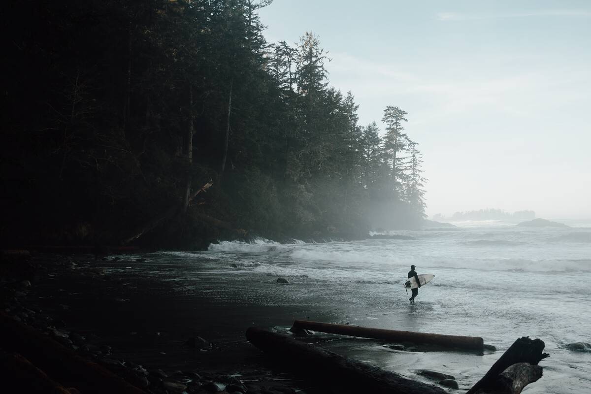 A surfer carrying a surfboard towards the water on a foggy day near Ucluelet BC. There is some driftwood in the foreground and evergreen trees in the background.