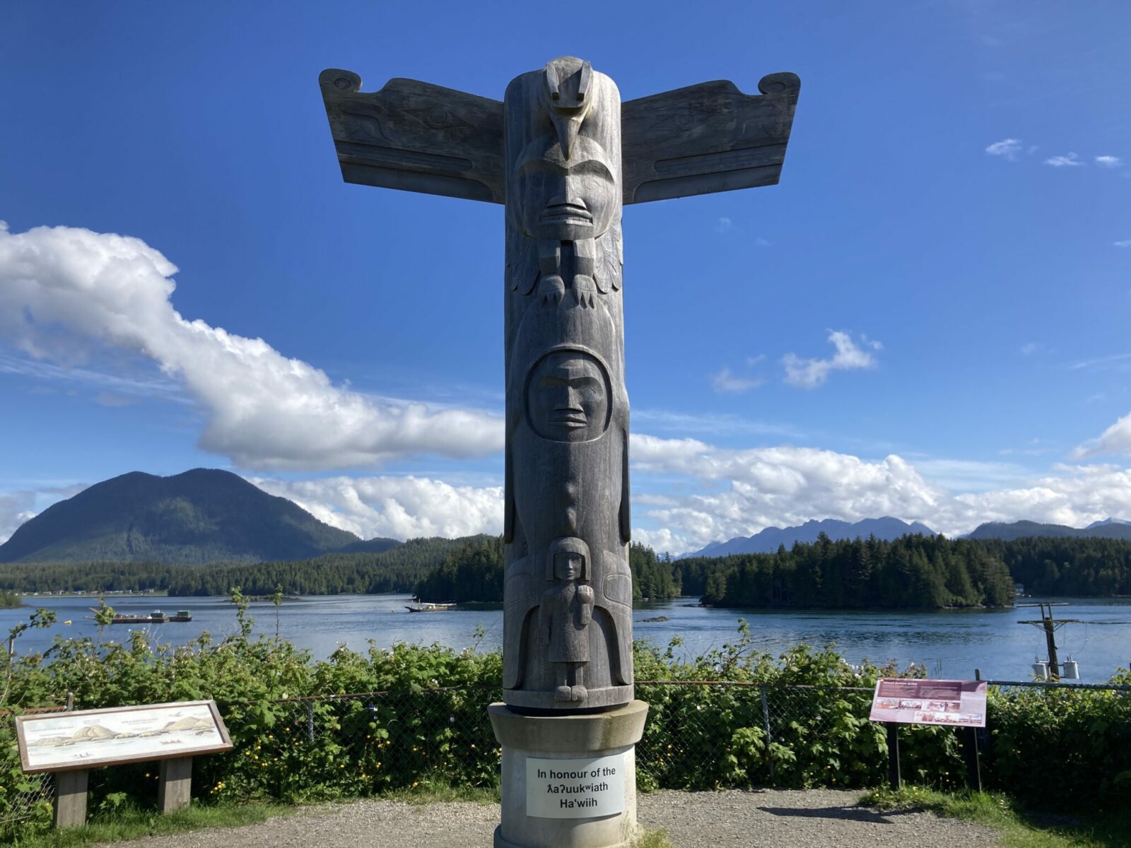 A wooden totem pole without paint in a city park at the waterfront. In the distance are forested islands and mountains on a mostly sunny day