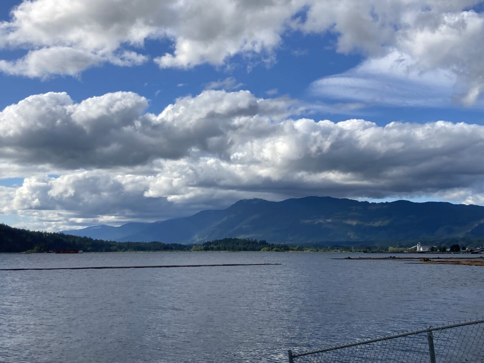 An inlet of the ocean in the foreground and the town of Port Alberni in the distance with a paper mill. There are forested mountains in the distance and the sky is partly cloudy