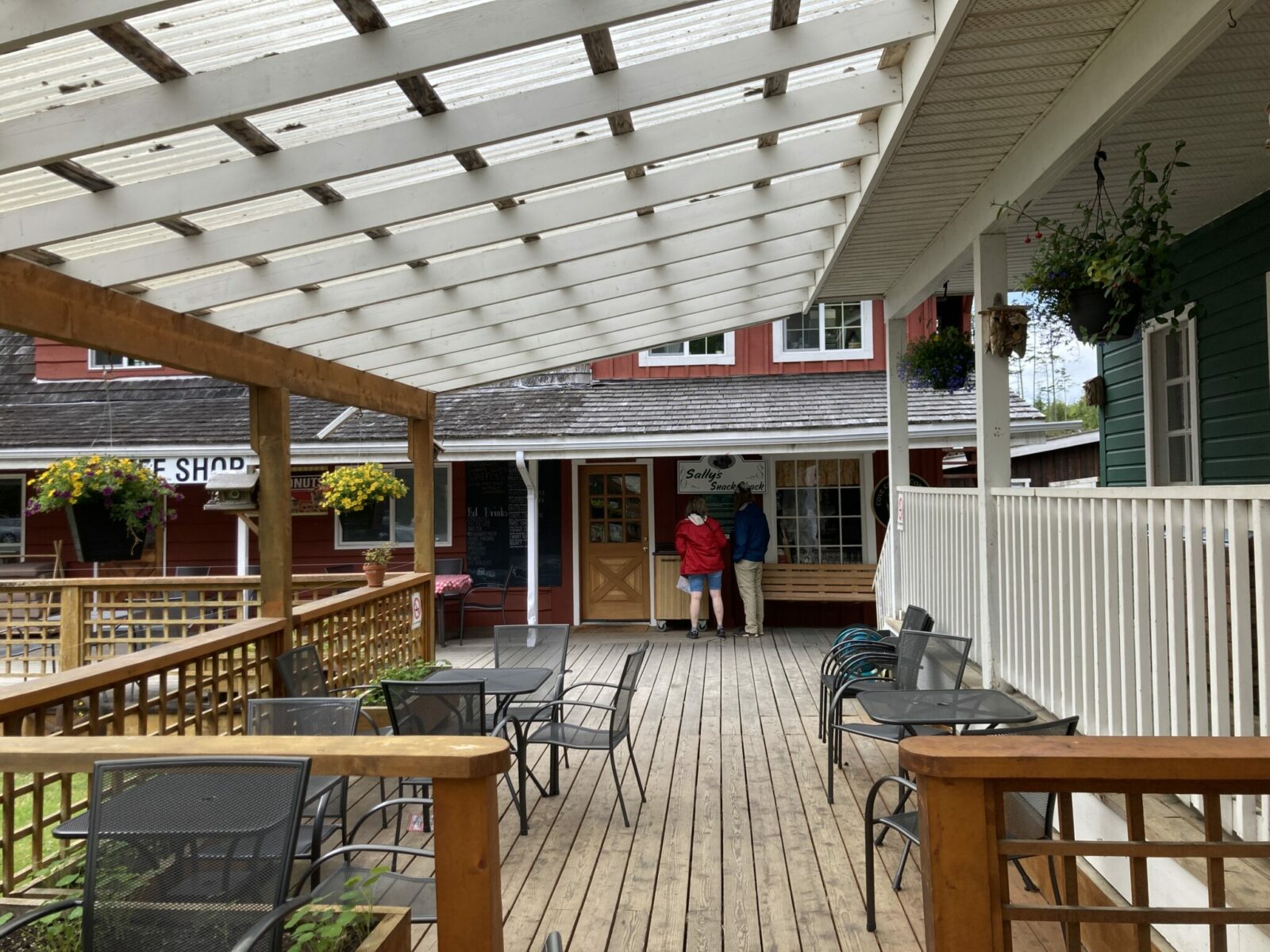 A wooden patio with a cover and tables and chairs. Two people are ordering coffee from the coffee bar in the distance.