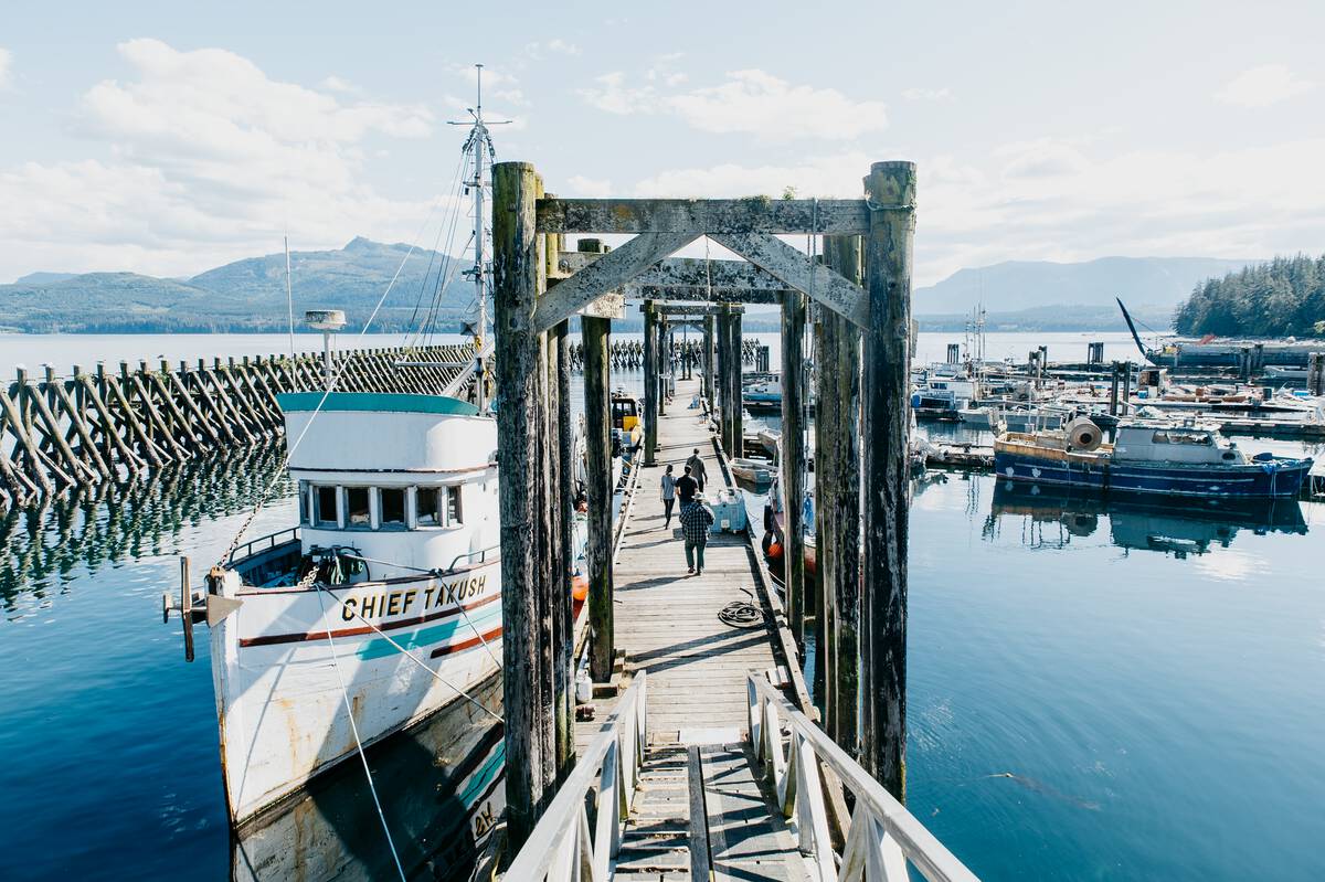 A marina with a wooden dock and many boats tied up to it. In the distance are forested hills and islands.