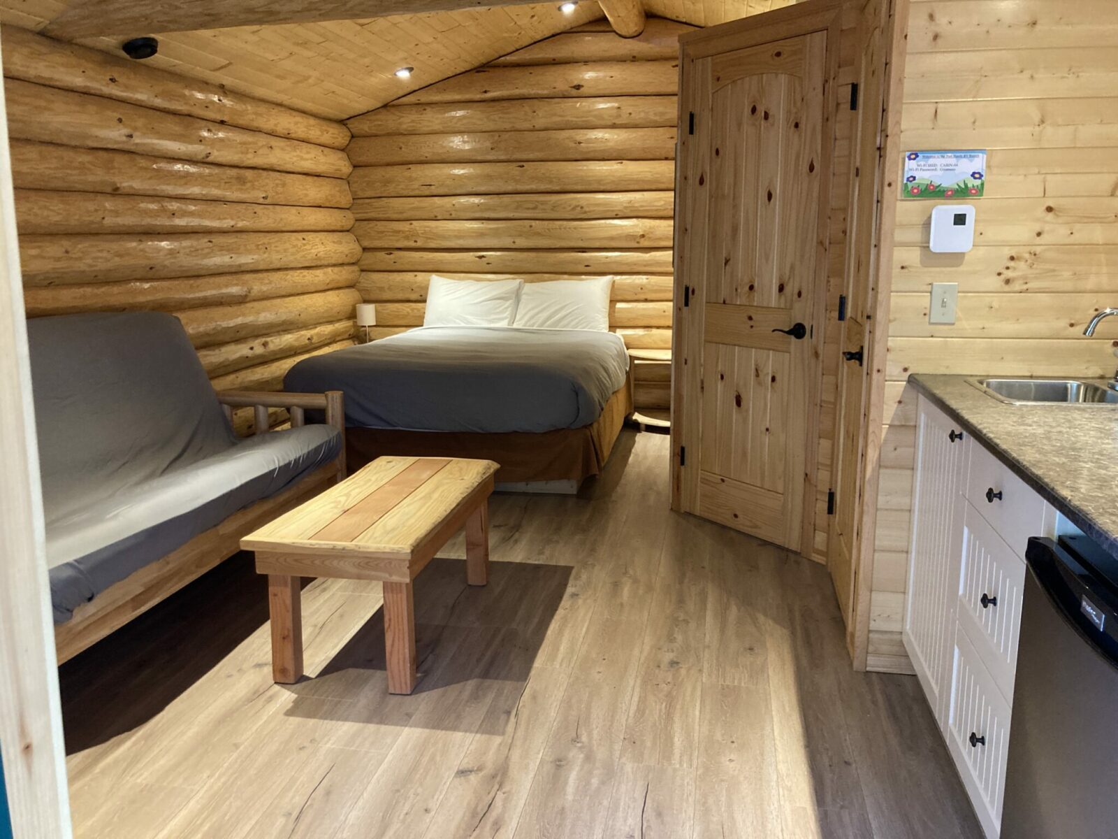 The interior of a small guest cabin. The walls are wood and there is a small kitchenette, a bed, a couch and a coffee table.