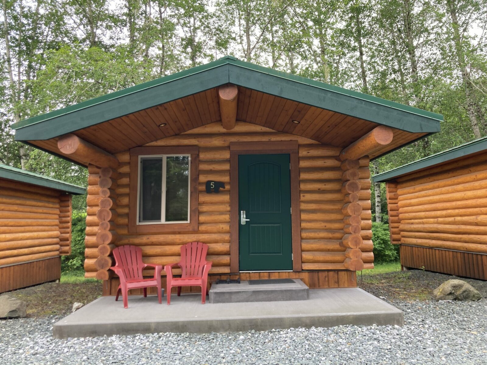 A small log cabin with a green metal roof. In front of the cabin is a concrete porch with two red chairs