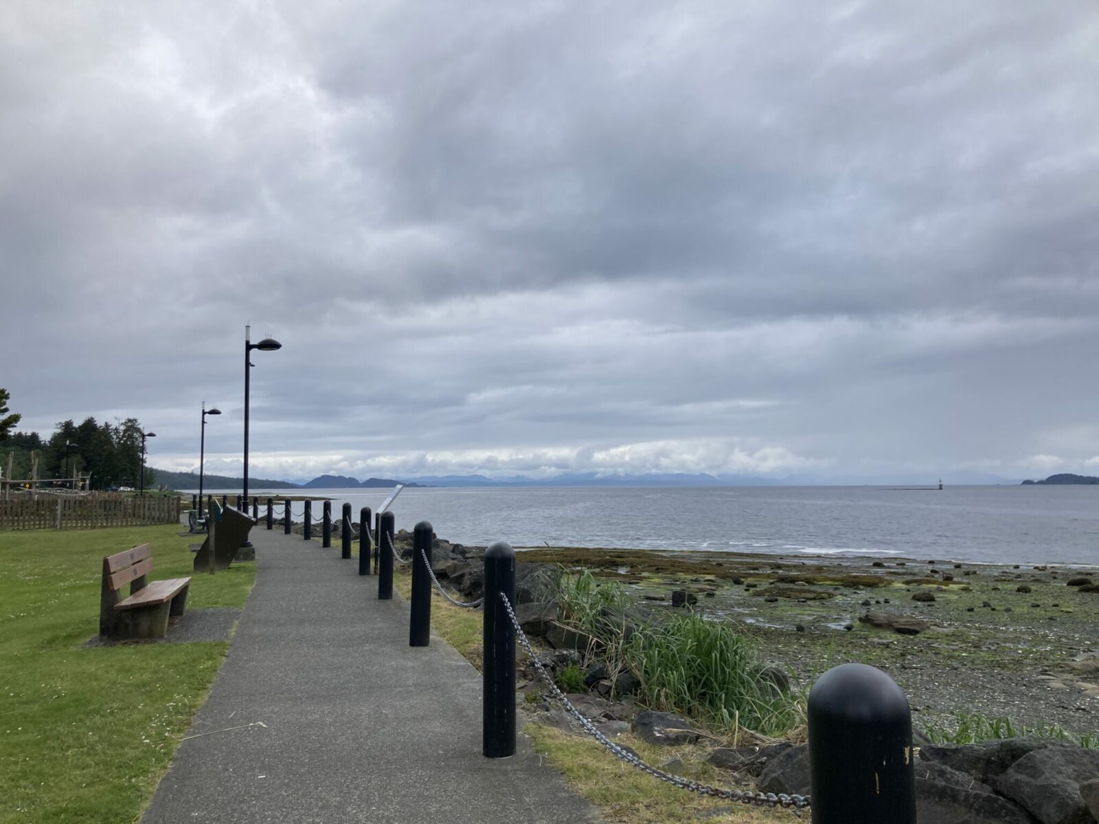 A paved walkway between the beach and a grassy field with beaches in Port Hardy. It is an overcast day with clouds letting a few distant mountains and hills peak through