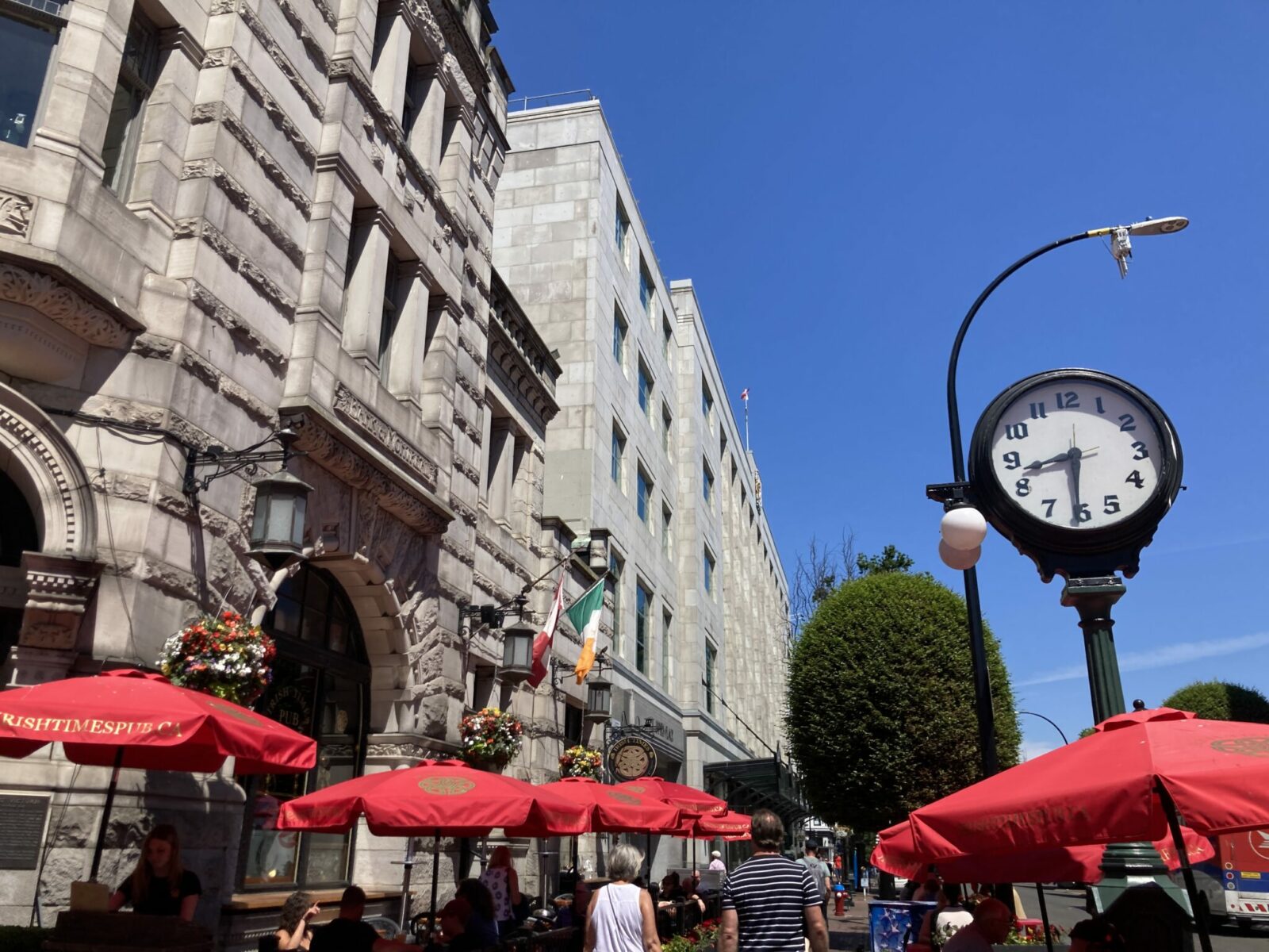 Stone buildings and an old clock on a city street on a sunny day. There are flags, hanging baskets and red umbrellas along the sidewalk.