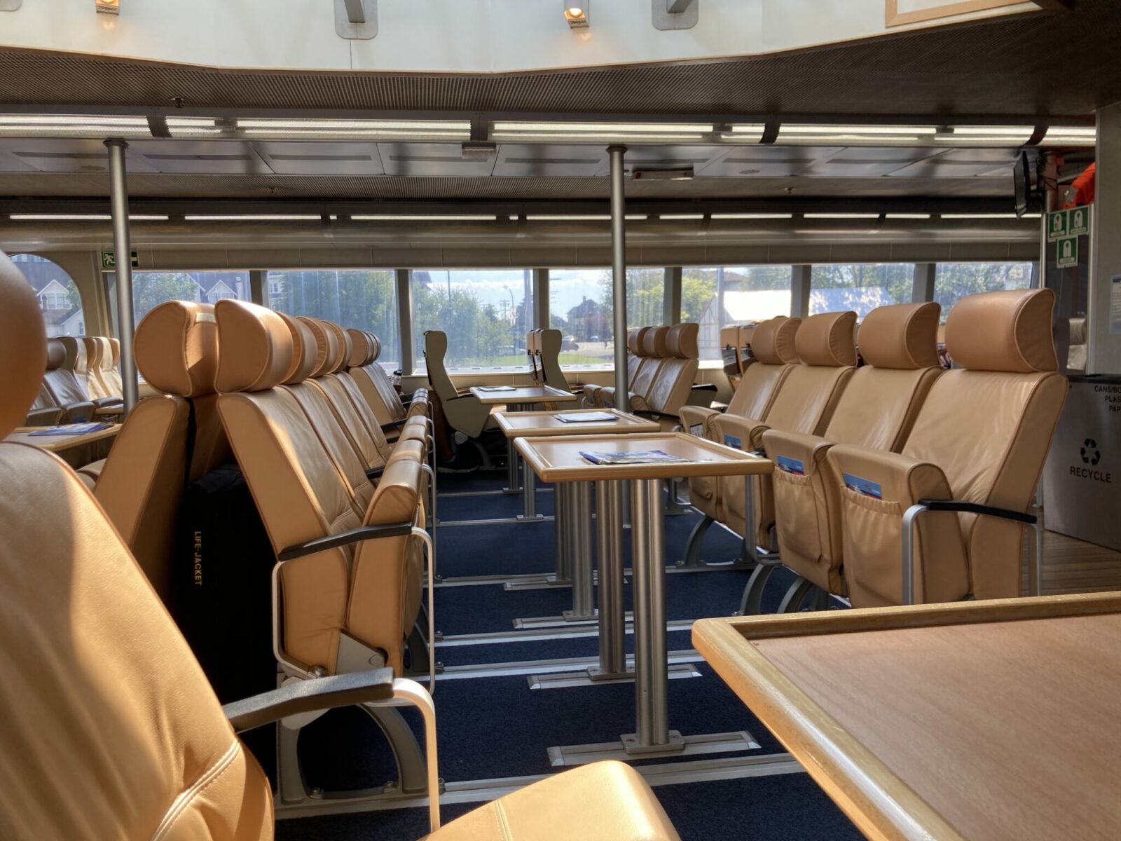 Seats on the upper deck of the Victoria Clipper. The seats are empty and facing each other at tables. Big windows let in the sunlight.