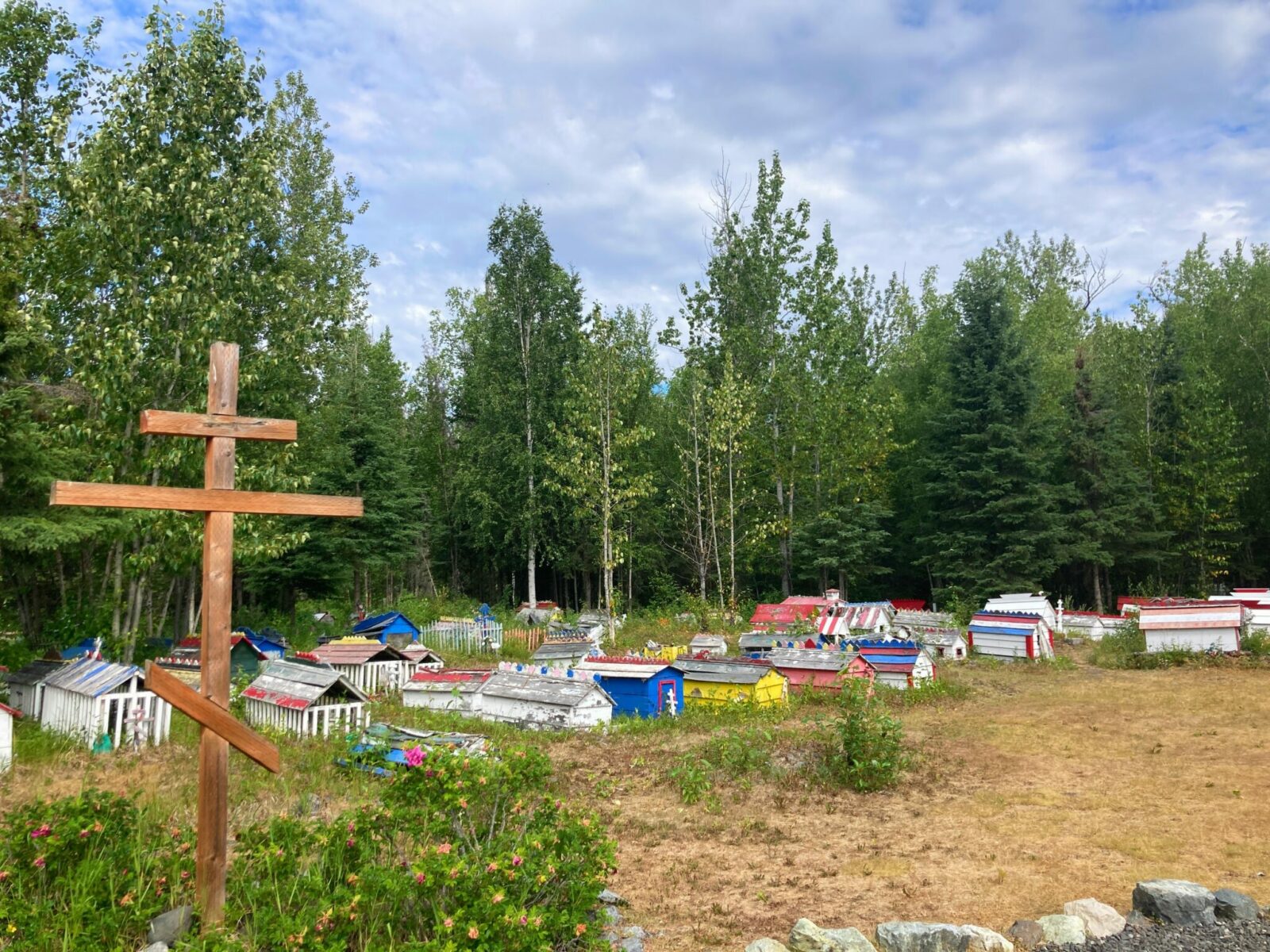 A russian orthodox cross in the foreground at the entrance to a cemetery in the forest. The cemetery has many small wooden structures called spirit houses to mark the graves. The spirit houses are brightly colored with paint.