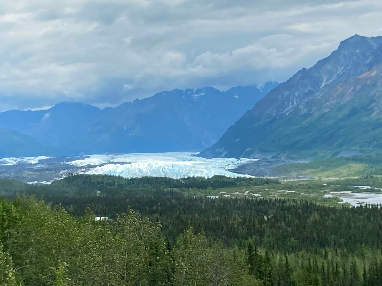 A glacier with the sun hitting blue and white ice in a valley between mountains with forest in the foreground