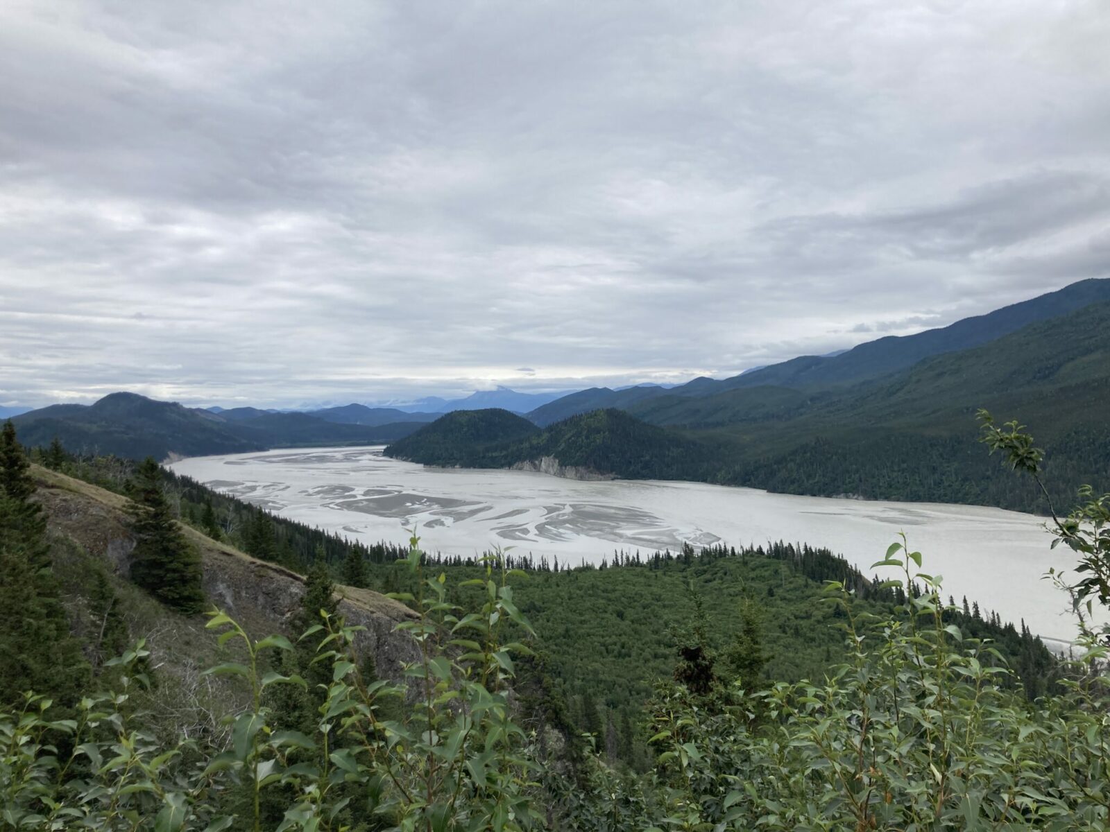 A view from above of the Copper River near Chitina, driving from Anchorage to McCarthy. The river is wide, braided and milky and has forest and mountains on both sides. Low clouds obscure the tops of the mountains