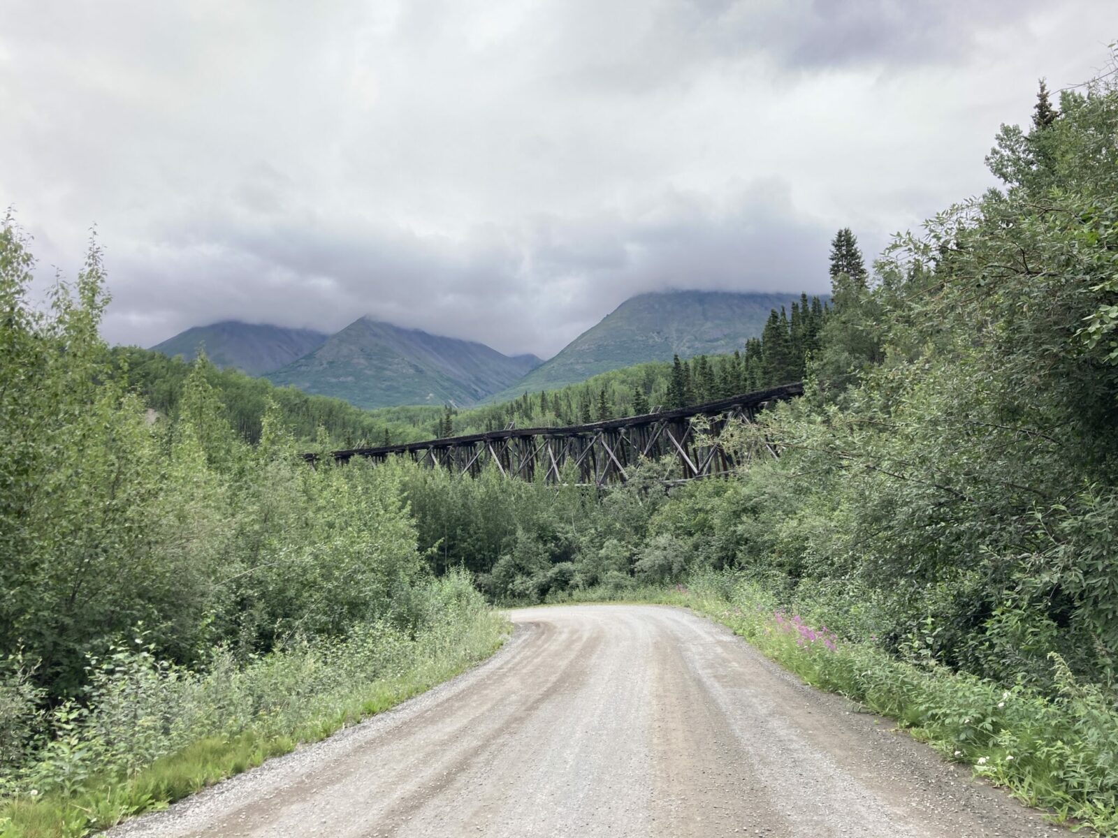 A historic partially collapsed railroad trestle from 1911 in the forest next to the gravel McCarthy Road. There are mountains covered in fog in the distance. The trestle is a highlight on the drive from Anchorage to McCarthy