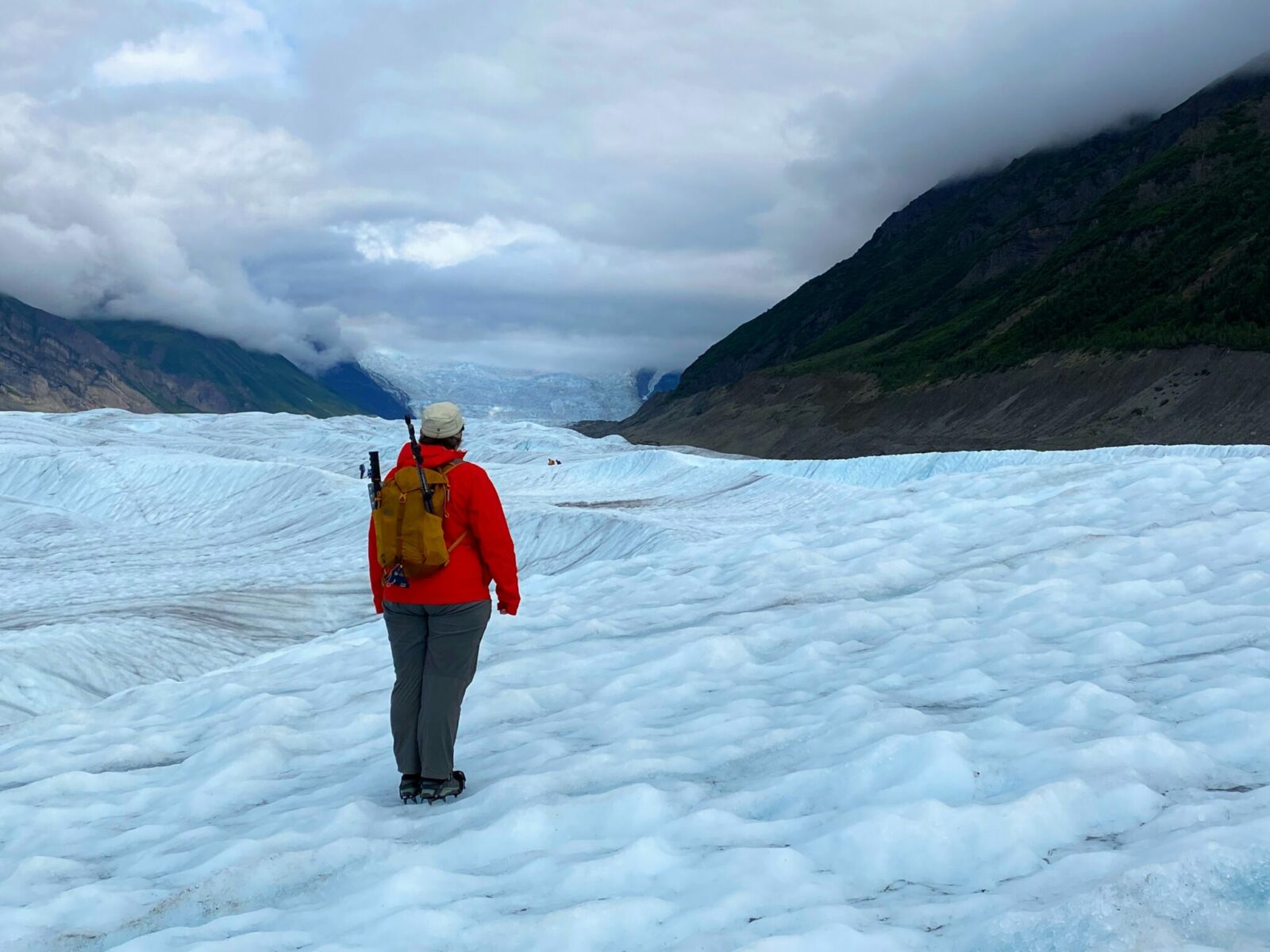 A hiker standing on the root glacier facing away from the camera. The hiker is wearing crampons, hiking pants, a red rain jacket, a beige hat and a yellow backpack