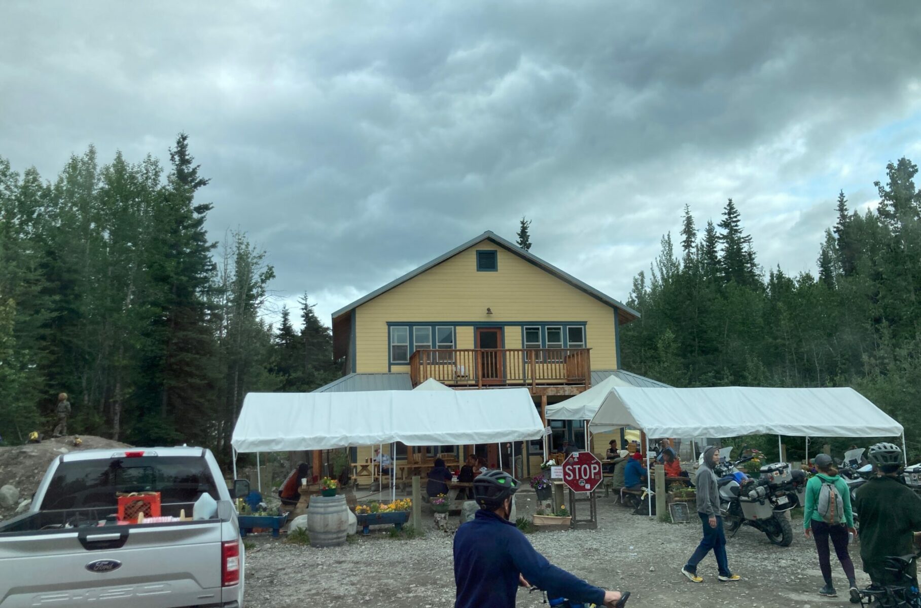 a yellow wooden building with picnic tables outside covered in white tents. A few people, bikes and a pick up truck are in the foreground near the building.