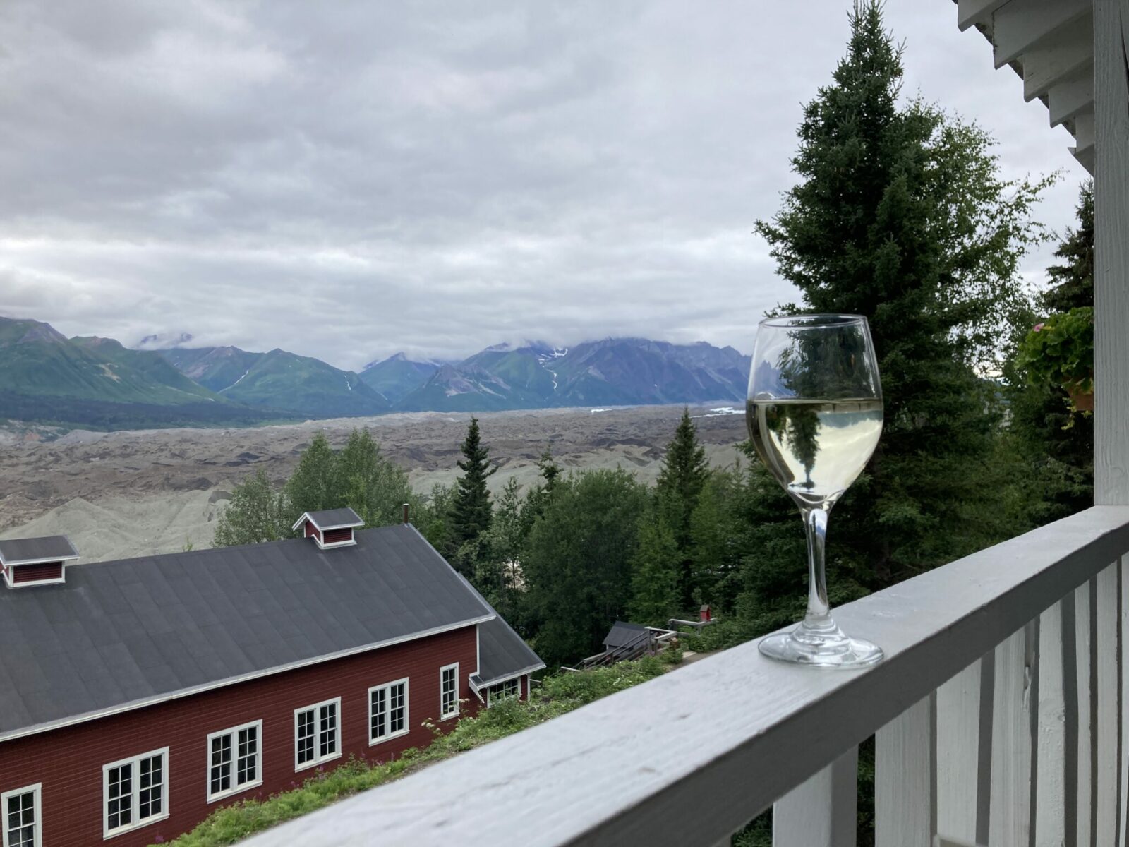 A glass of white wine on a handrail. Beyond the porch there is a view of a red and white building, trees, and the gravel covered Kennicott Glacier and mountains of Wrangell-St Elias national park.