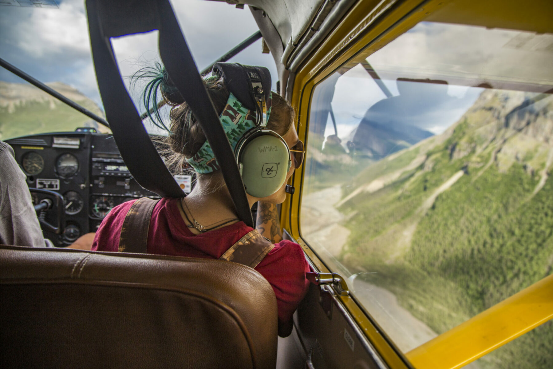 A person looking out the window of a small plane at mountains below. The person is in the co pilot seat and wearing a headset. 