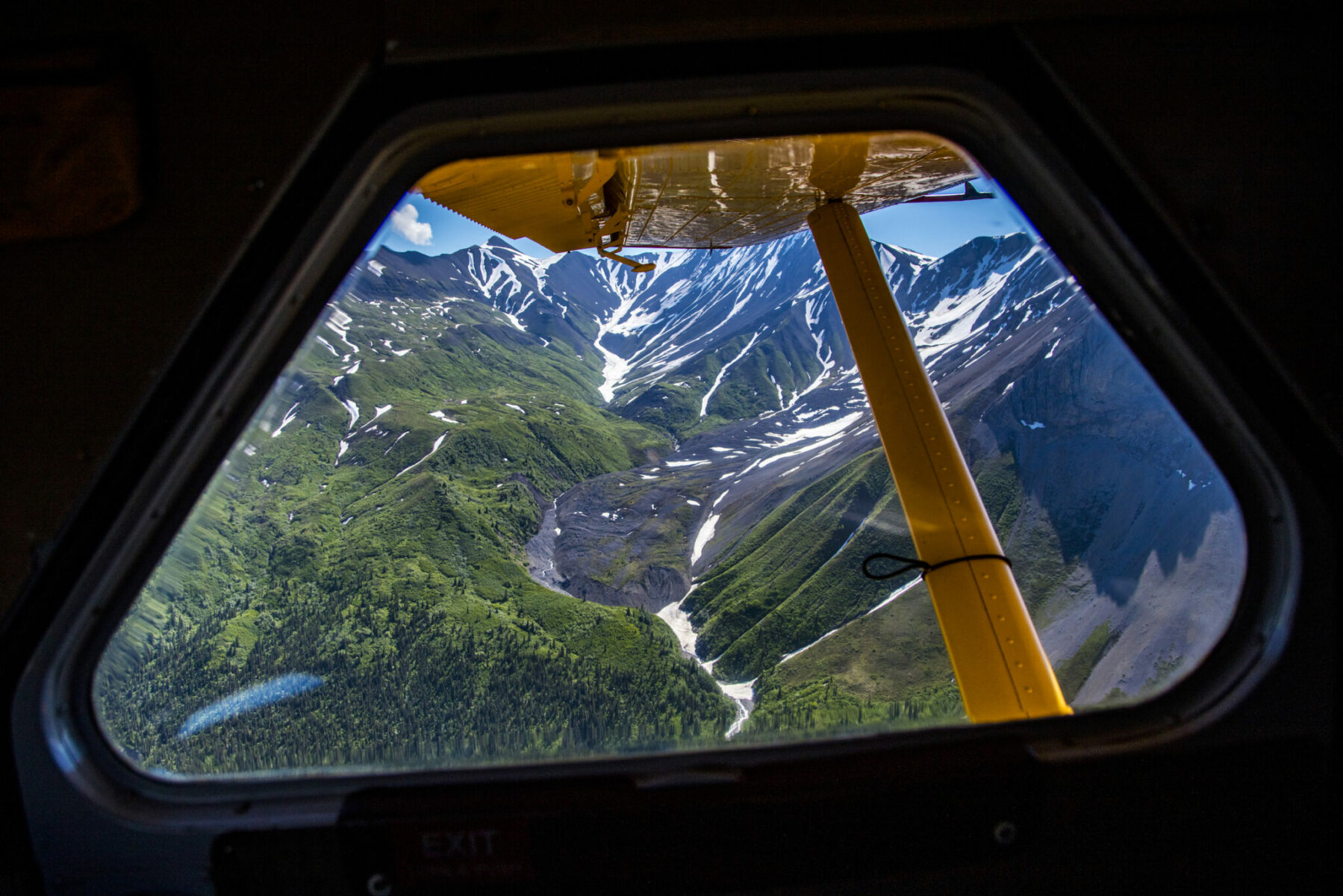 A small plane airplane window with a view of the wing and snow covered mountains and forest in the background