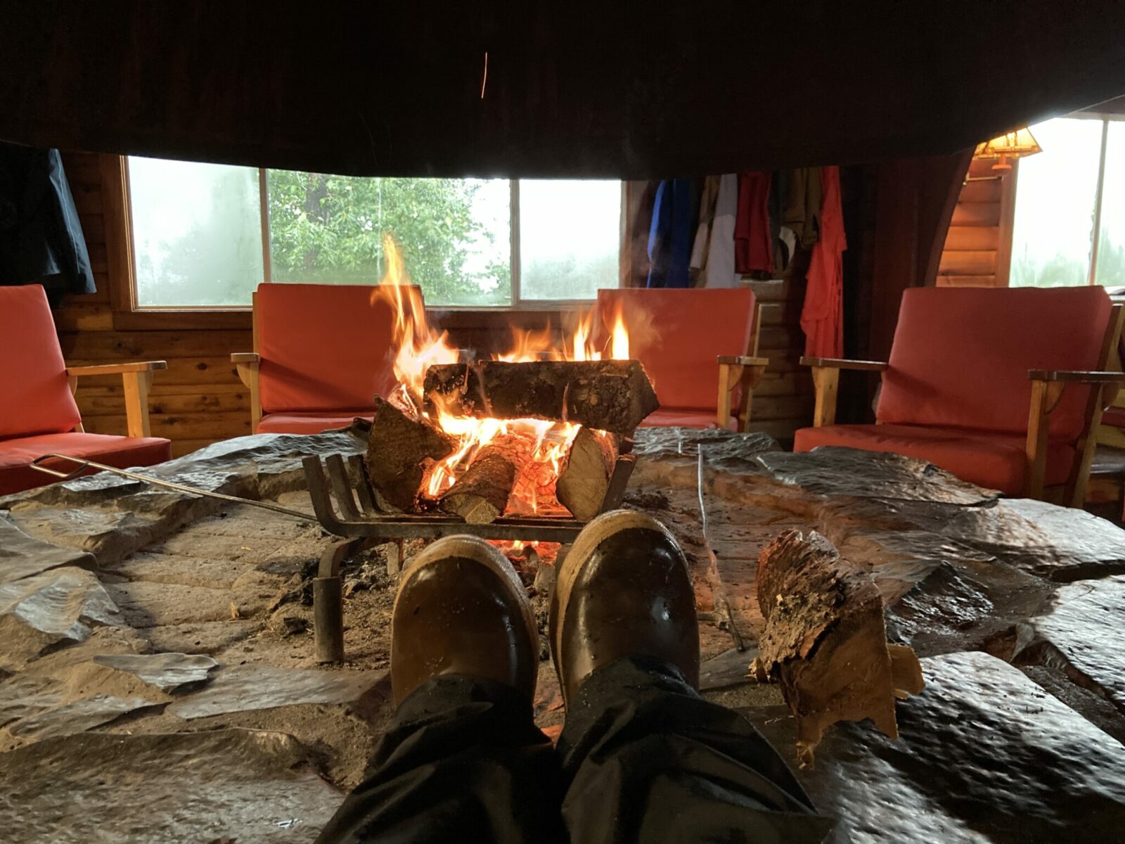 A person's rubber boots and rain pants sitting on the edge of a fireplace to dry with a roaring fire and chairs around the firepit at Brooks Lodge in Katmai National Park