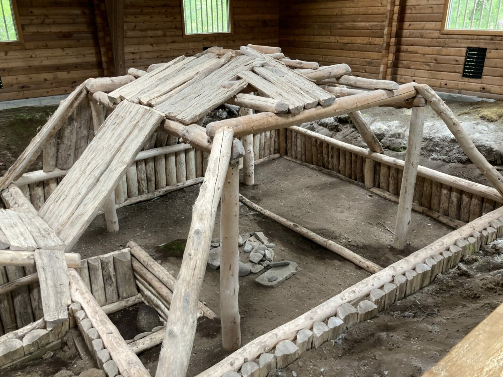 A recreation of an archaeology site that is a partially below ground house with logs above the ground. The site is inside of a building and has a dirt floor.