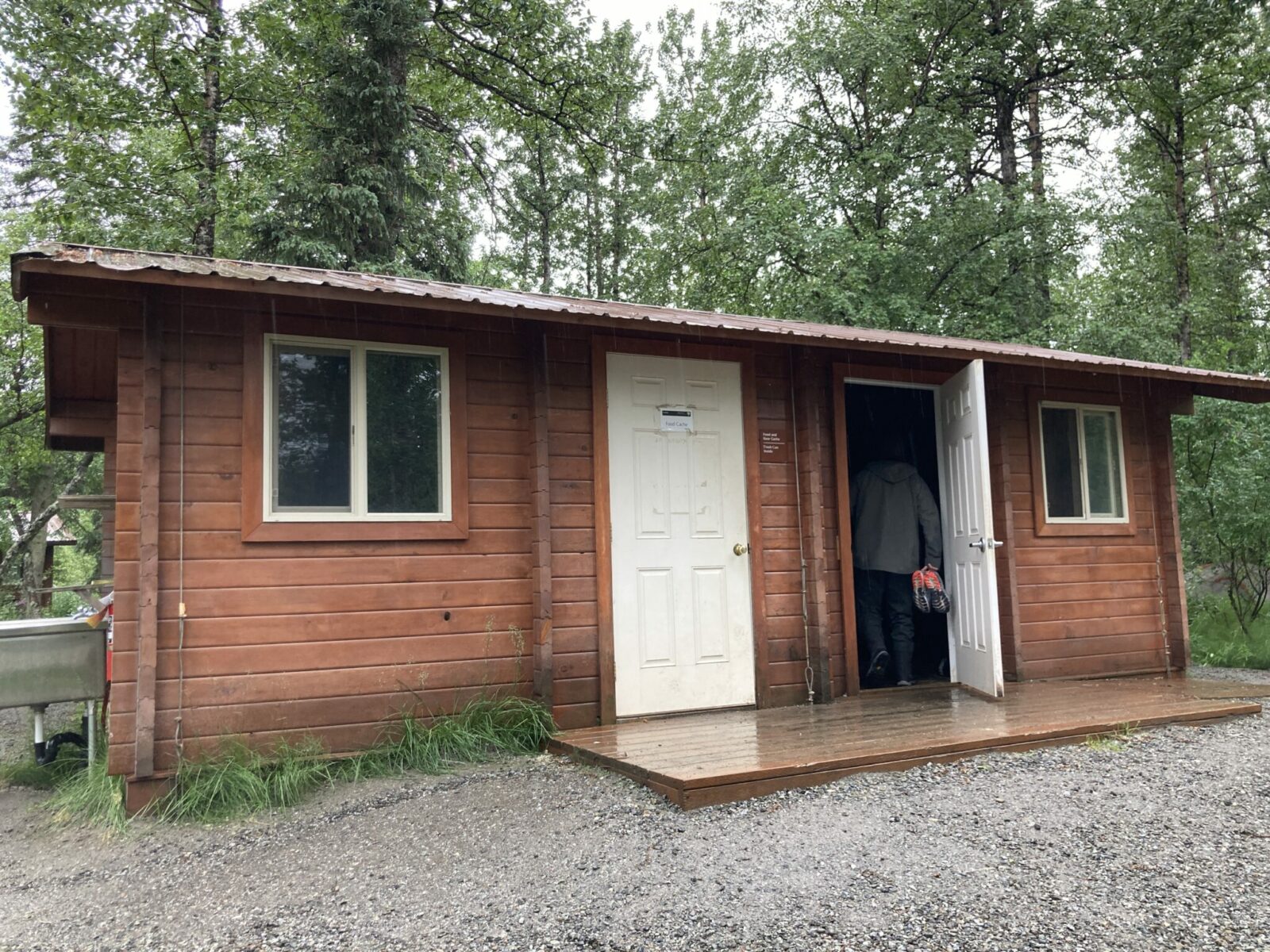 A wooden building with a small deck in front, surrounded by gravel in front and forest around it. There are two doors and two windows and a metal roof. A person is walking into the door on the left