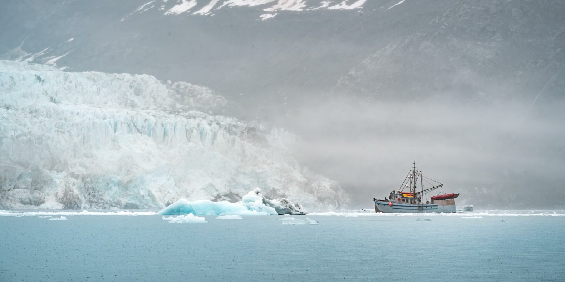 A glacier in the fog with ice bergs in front of it. A fishing boat is near the glacier and the ice bergs