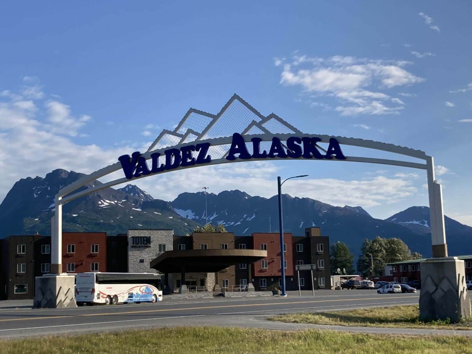 A white metal sign that says Valdez Alaska in purple letters over the highway. In the background are snow capped mountains and a new hotel with brown and red paint.