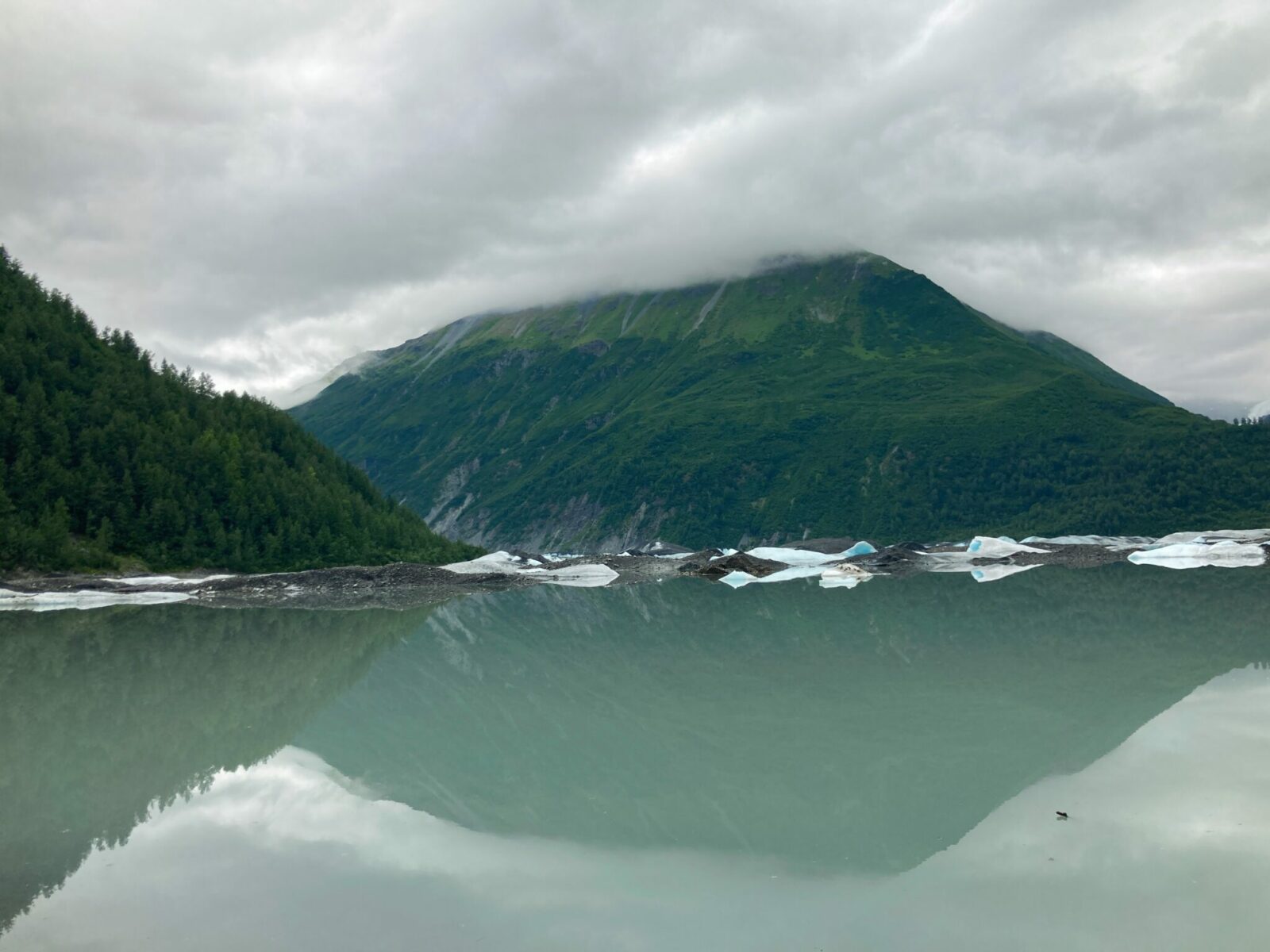 Valdez Glacier Lake near Valdez Alaska. it is an overcast day and there are forested hillsides surrounding a silty and milky lake below a glacier that is not visible. There are icebergs that are blue and white floating on the water and on the beach.