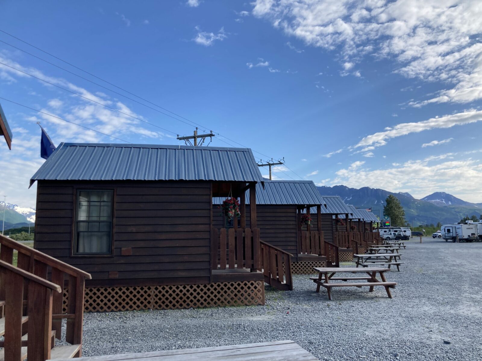 A series of small, wooden cabins with metal roofs in a gravel parking lot at an RV park in Valdez, Alaska. Each cabins has a flower basket, a small porch and a picnic table in front. There are mountains in the distance