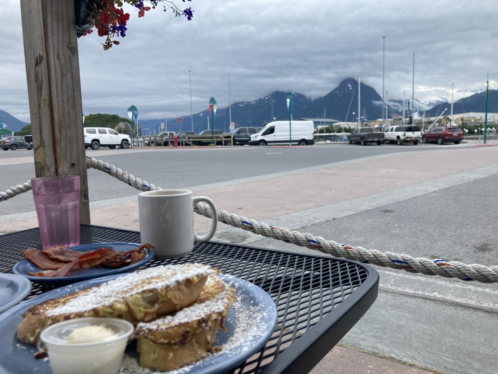 A black cafe table on the patio of the Fat Mermaid Restaurant in Valdez, Alaska. There is french toast, bacon and coffee on the table and in the distance is the parking area for the harbor, the harbor and distant mountains across the bay.