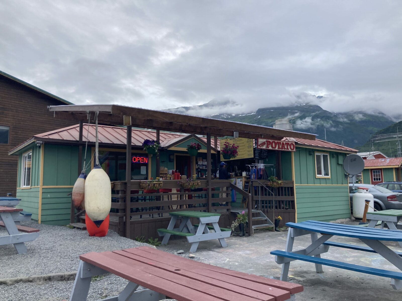 A small green wooden building with an open sign that says "the potato", the name of the restaurant. There are a few tables on the porch and picnic tables around the building.