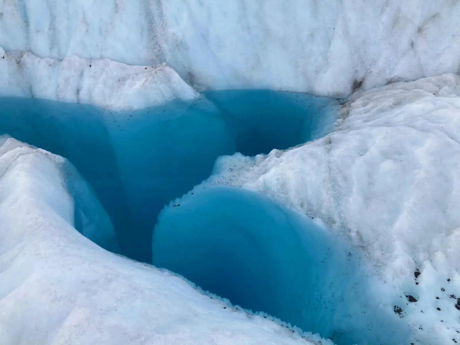 A deep, blue pool of water on the surface of the Root glacier. The water is clear but you can't see the bottom. The pool is lined with white ice and some gravel frozen into it.