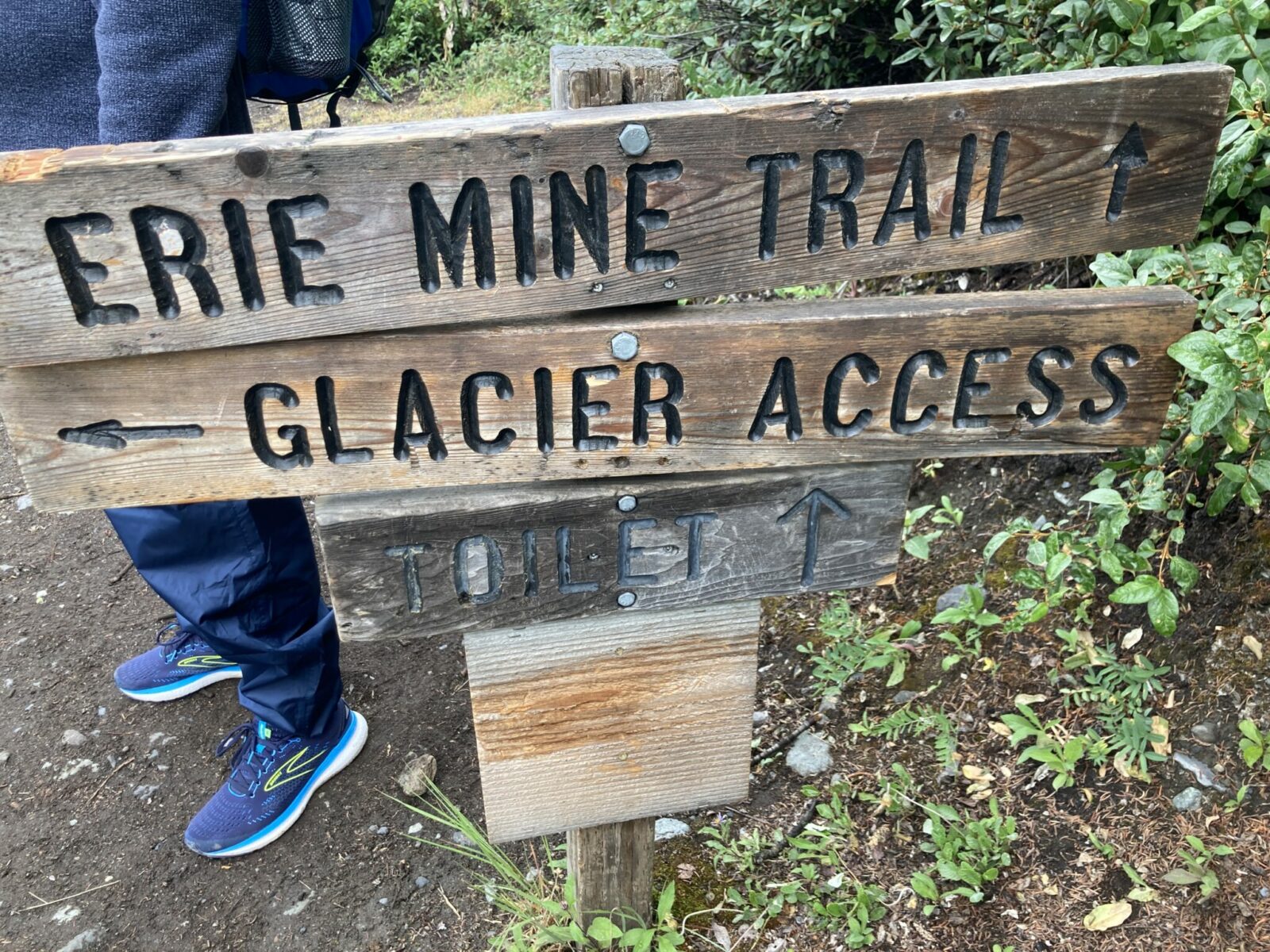 A wooden trail sign that says Erie Mine Trail with an arrow pointing straight ahead, and one saying glacier access with an arrow to the right. There is a smaller sign that says toilet with an arrow pointing ahead. A hikers feet are visible behind the sign.