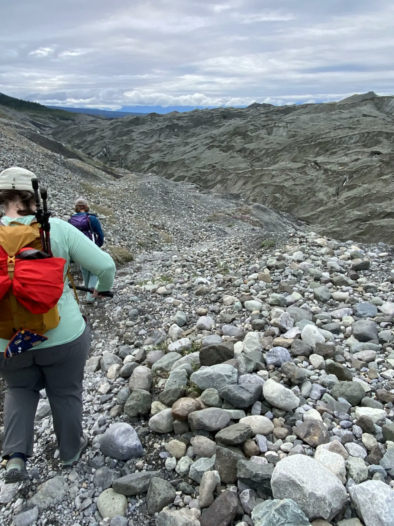 A gravel hillside with two hikers walking down a steep slope towards a glacier