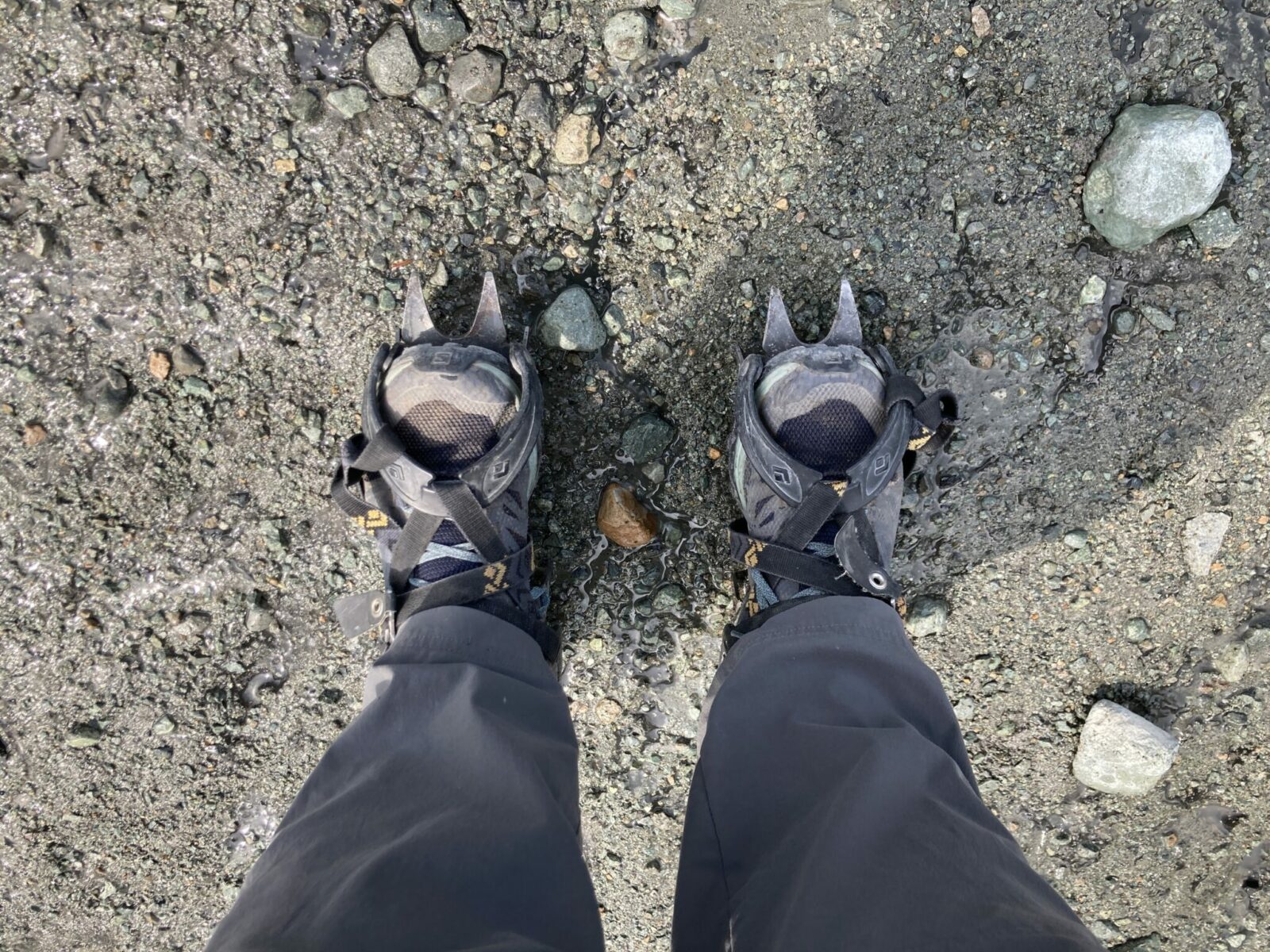 A person's feet wearing hiking pants, hiking shoes and crampons on the gravel edge of Root Glacier