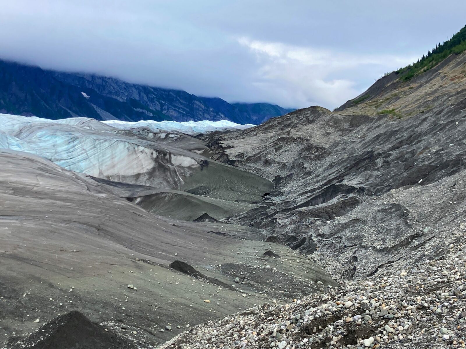 The edge of the blue Root Glacier where it meets the graveled edge of the hillside next to it. There is a transition zone of gravel and rock between the glacier and the hill. Mountains obscured by clouds are in the background