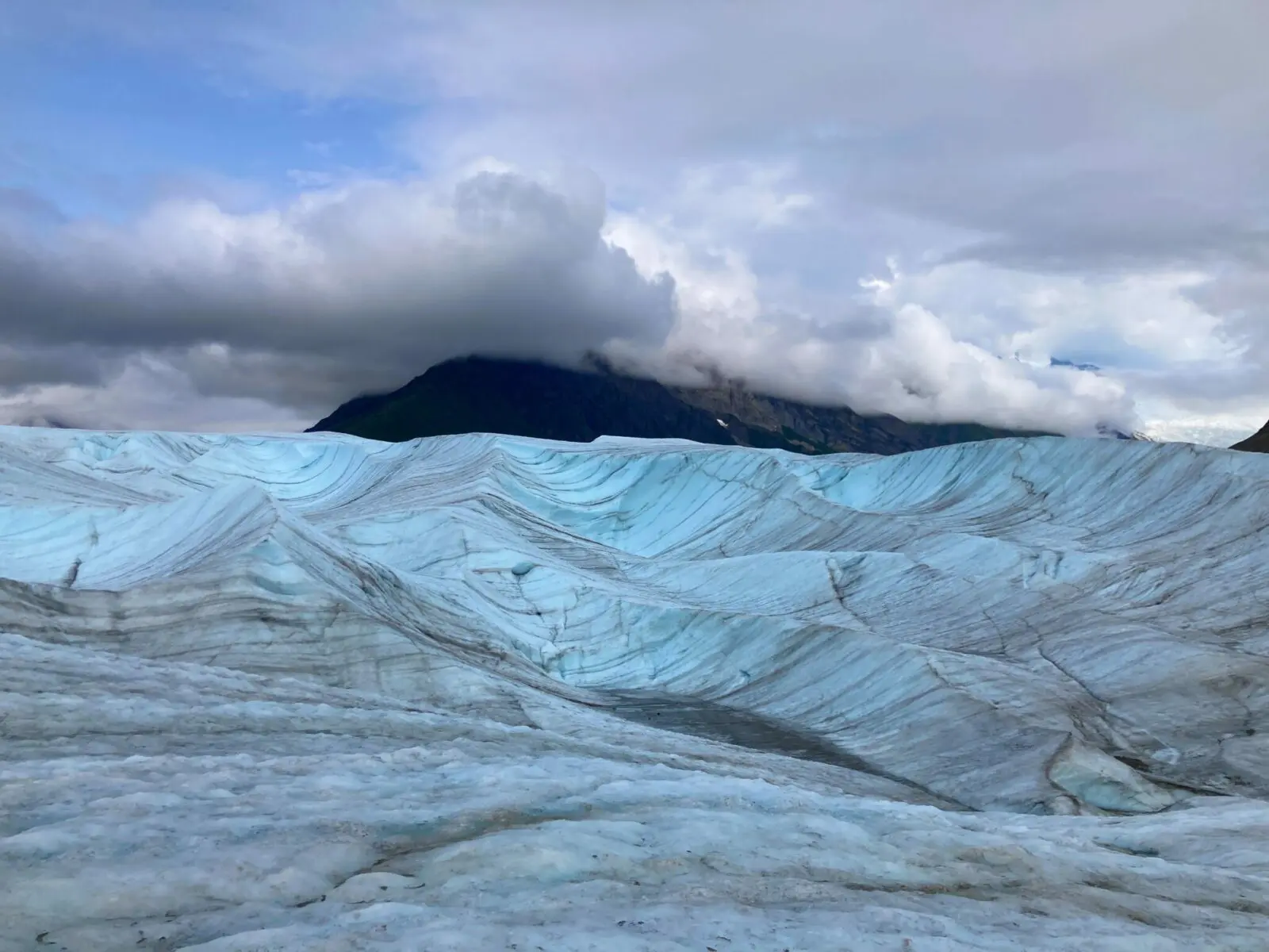 On top of the Root Glacier, there are streaks of grey in the blue ice. The ice has mounds like small hills blocking the distant views. One mountain is partially visible behind the glacier in the clouds