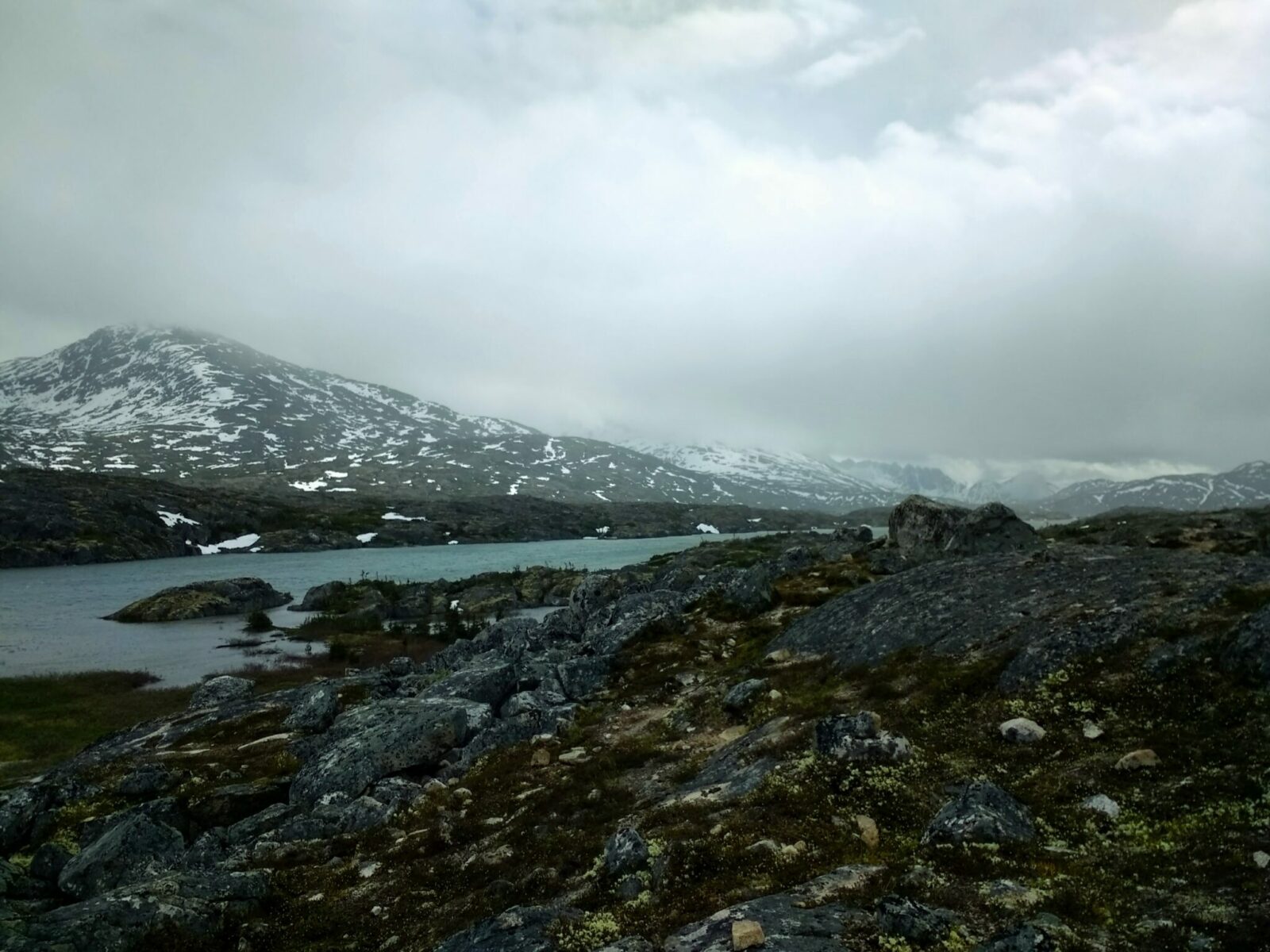 A rocky alpine area with a greenish lake and some mountains with a bit of snow. You can only see the bottom of the mountains as thick gray clouds are hanging low over them