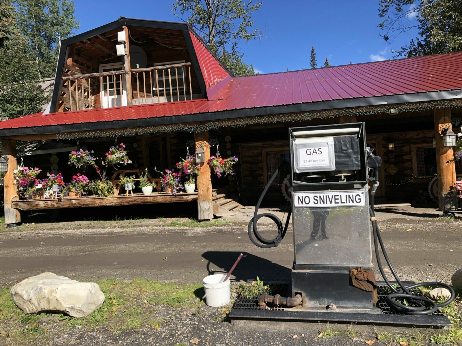 A wooden roadside lodge with a red metal roof and lots of flower baskets with a single old school gas pump in front.