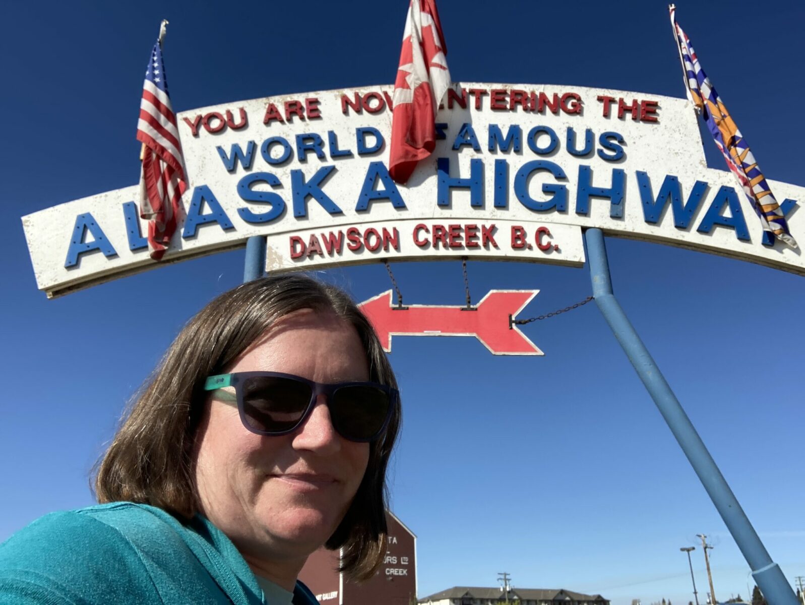 A person smiling and wearing sunglasses standing under the Alaska Highway milepost 0 sign.