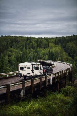 A black pick up truck pulling a camper trailer on a curved wooden bridge on an overcast day through the forest.