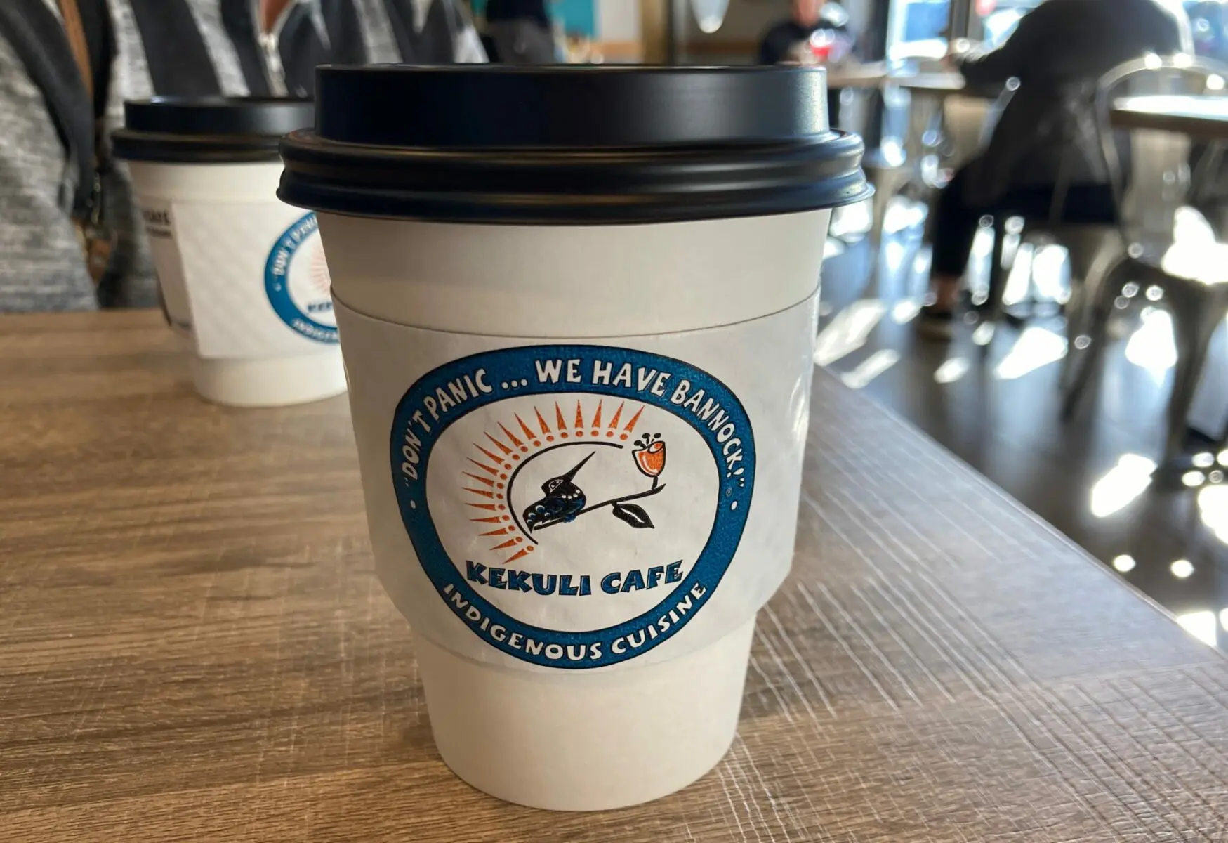 A white coffee cup with a black lid on a wooden table in a coffee shop. The cup is labeled Kekuli Cafe Indigenous Cuisine