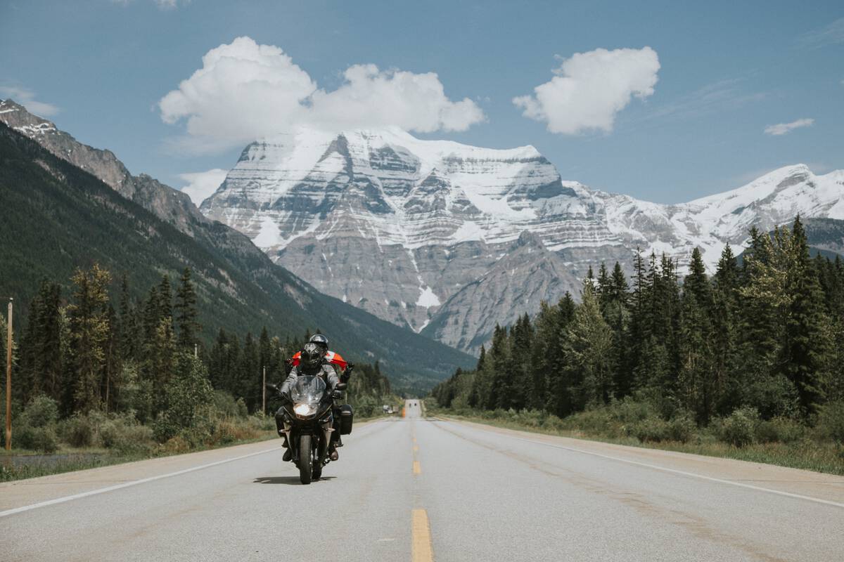 A high snowy mountain above a forested highway. The mountain has clear layers of rock showing up the side