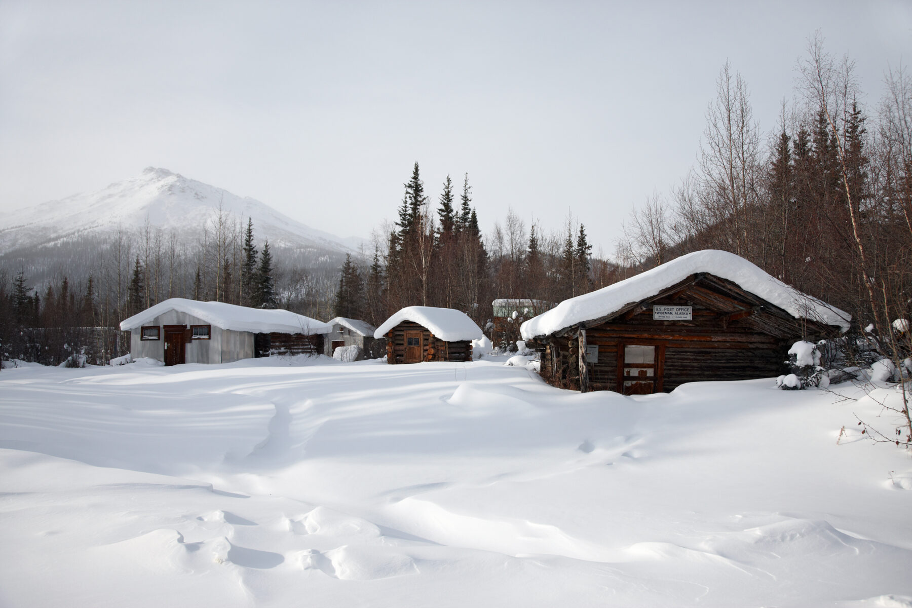 Deep snow covers a cluster of small log cabins with a few trees and mountains in the distance